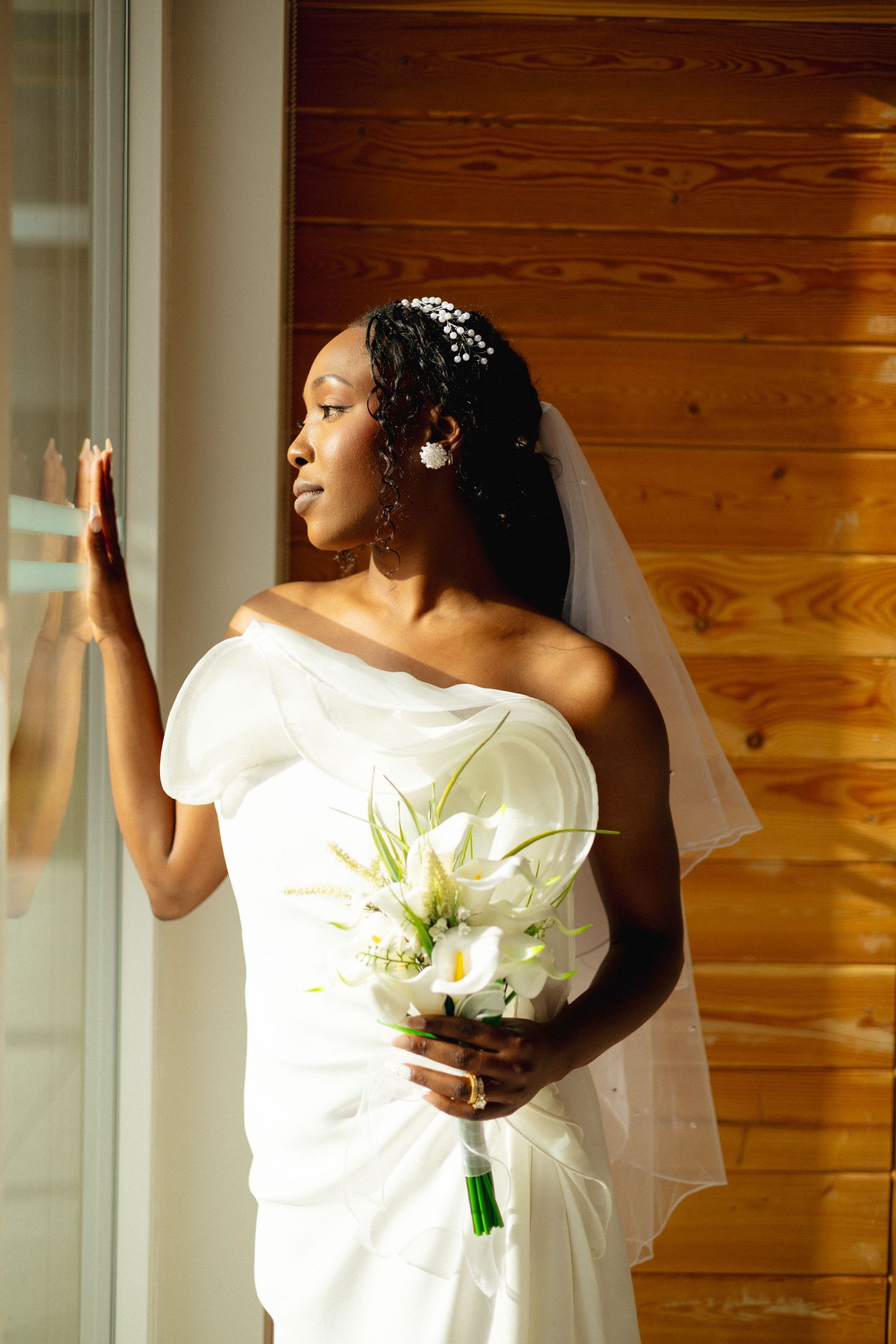Elegant Black bride in strapless white gown holds calla lily bouquet while gazing out sunlit window.