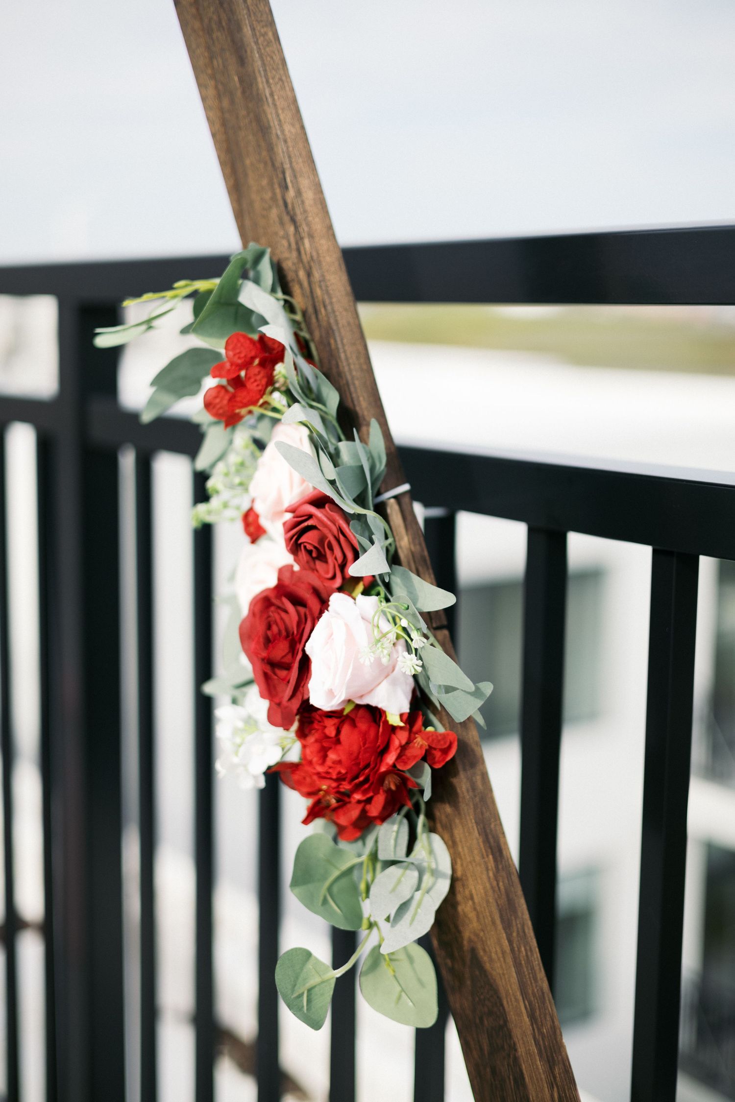 Red and white roses with eucalyptus greenery arranged on a rustic wooden wedding arch with black metal railing.