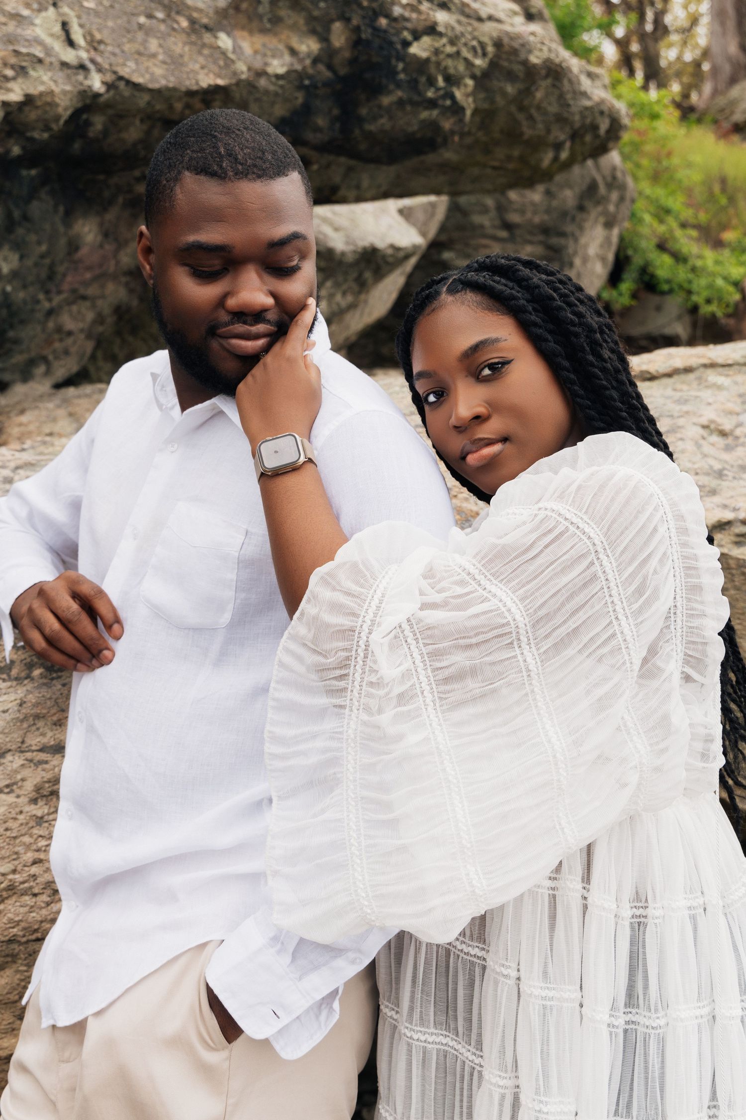 A couple in white outfits poses against rocks, woman touching man's face tenderly.