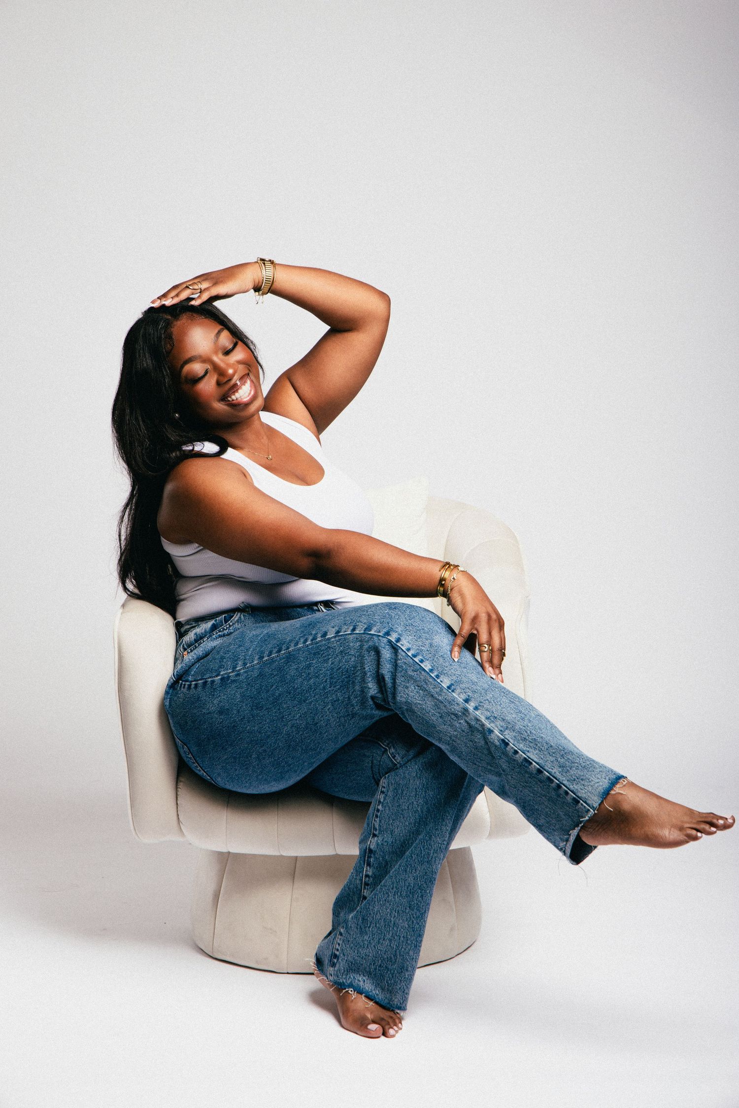 A smiling woman in white tank top and blue jeans sits relaxed in a white chair, hand raised to her head.