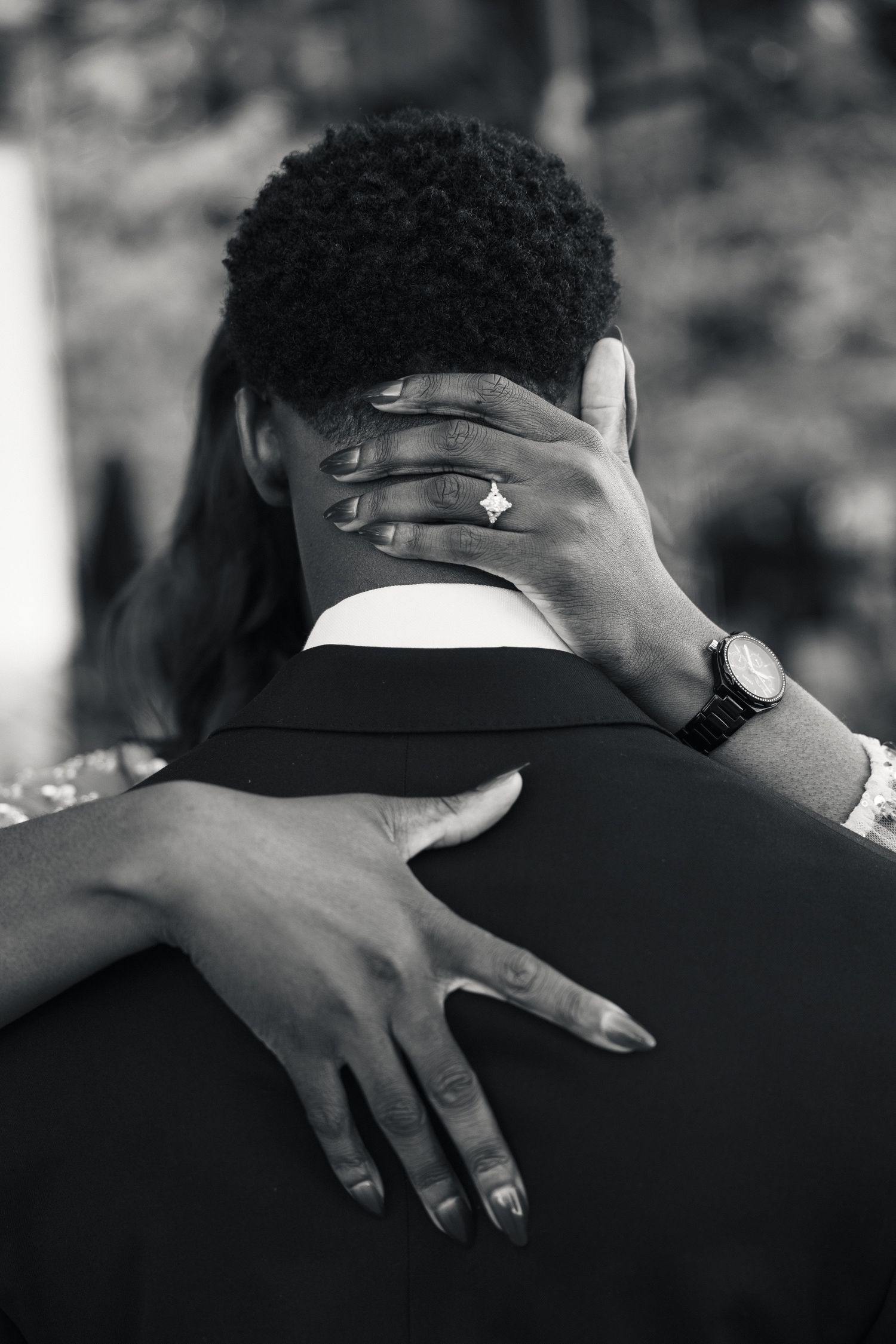 Black and white photo of a couple embracing, woman's hand with diamond engagement ring resting on man's neck.