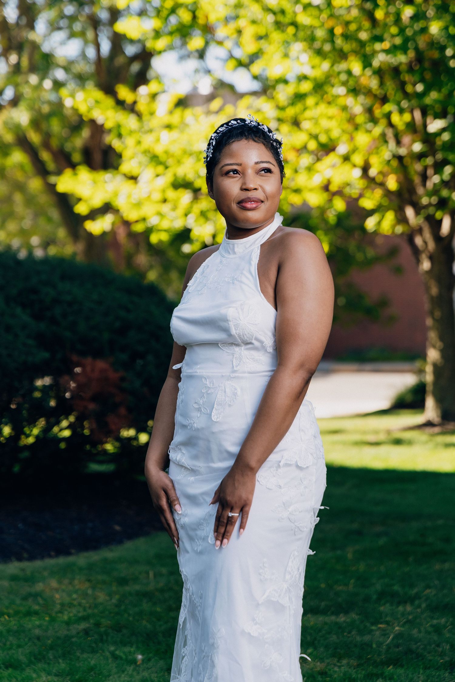 Elegant bride wearing white halter wedding dress with embroidery stands in sunlit garden setting.