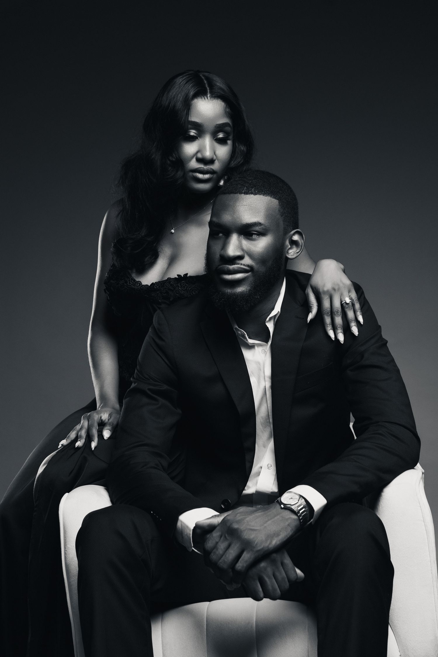 Elegant black and white portrait of a couple in formal attire, woman standing behind seated man in studio setting.