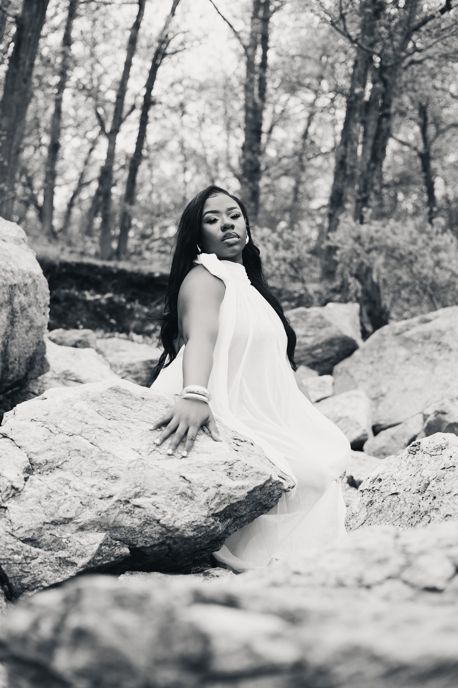 Black and white photo of a woman in a white dress posing gracefully on rocks in a forest setting.
