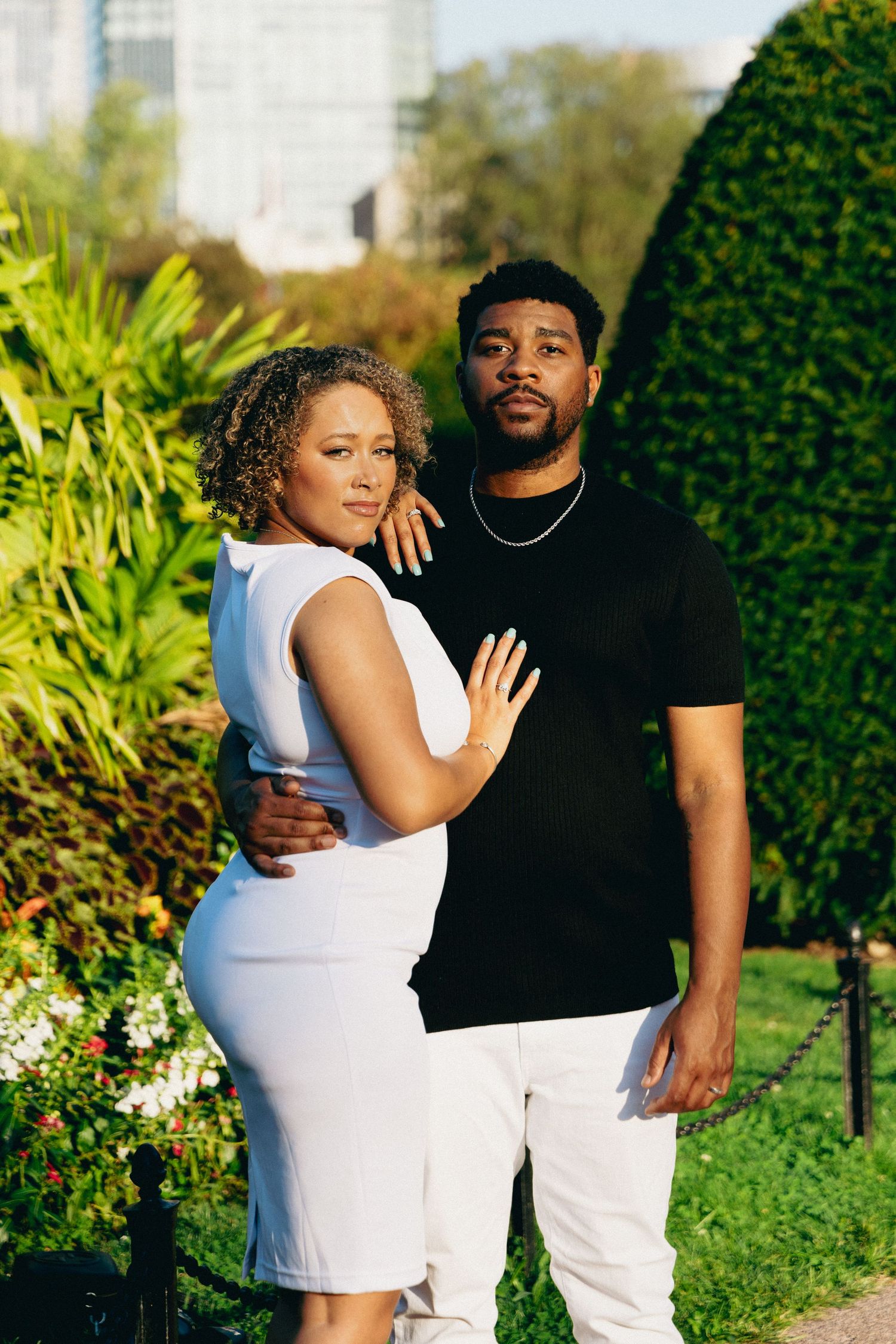 A stylish couple poses in a lush garden, she in a white dress and he in a black shirt and white pants.