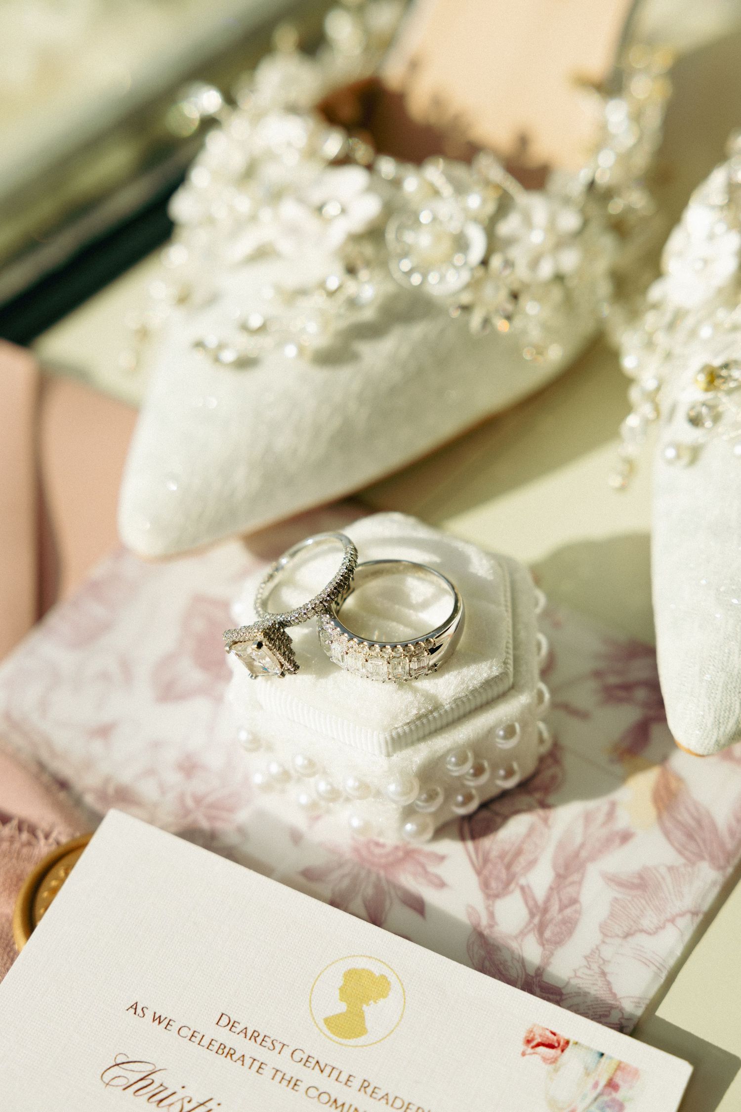 Elegant bridal details featuring diamond wedding rings on a pearl ring holder beside white lace heels and a floral wedding invitation.