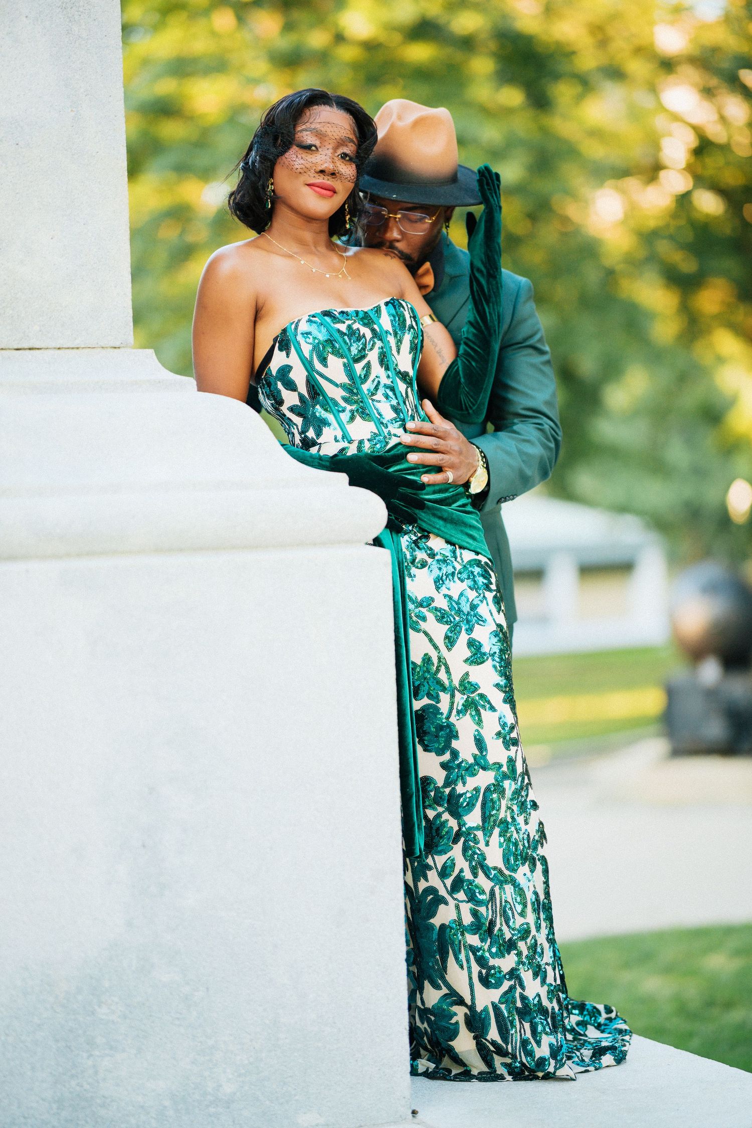 Couple posing outdoors, woman in green floral gown with gloves, man in green suit embracing her by a stone column.