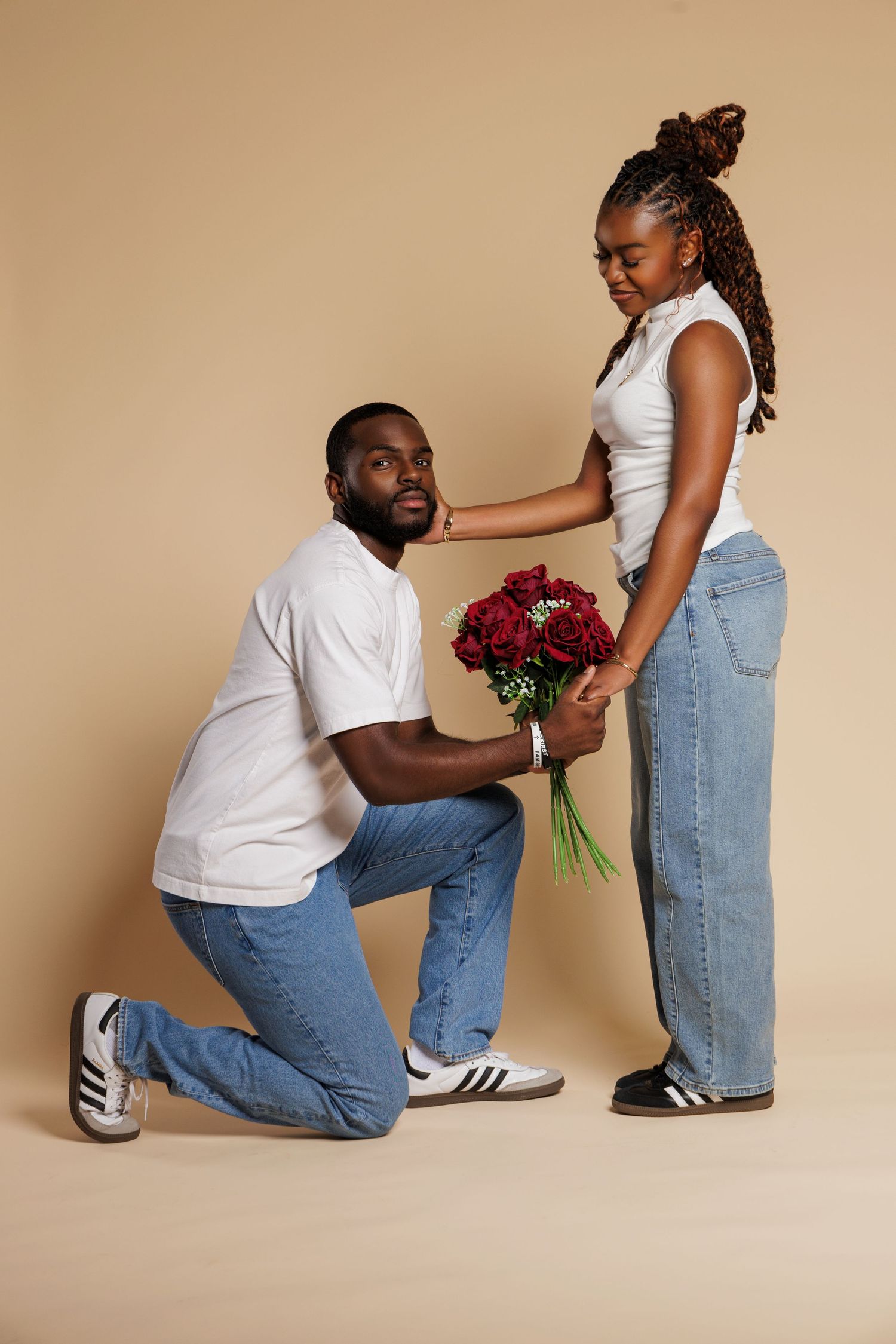 Man kneeling on one knee presenting red roses bouquet to woman in white top and jeans on beige background.