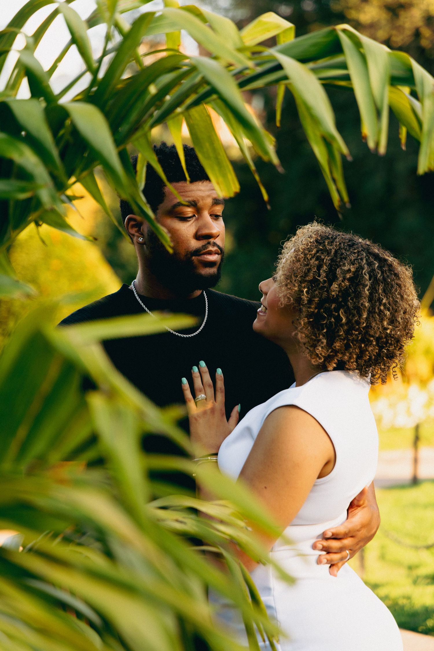 A couple shares a romantic moment surrounded by lush tropical greenery, the woman in white gazing up at her partner.