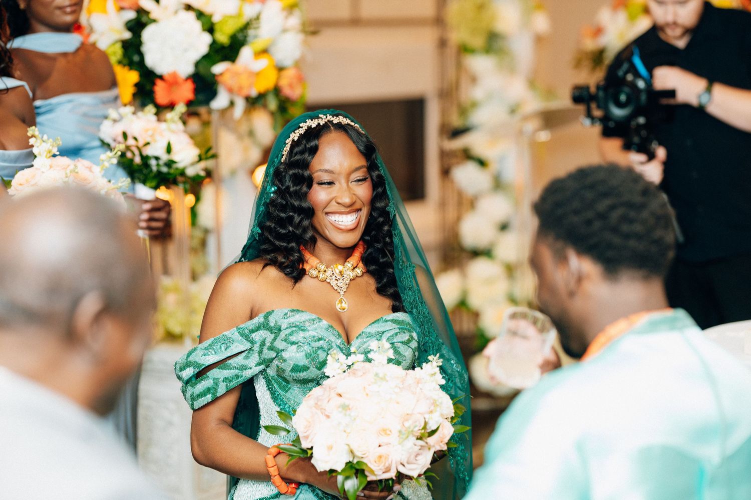 Smiling bride in green off-shoulder gown and teal veil holds white bouquet at colorful wedding ceremony.