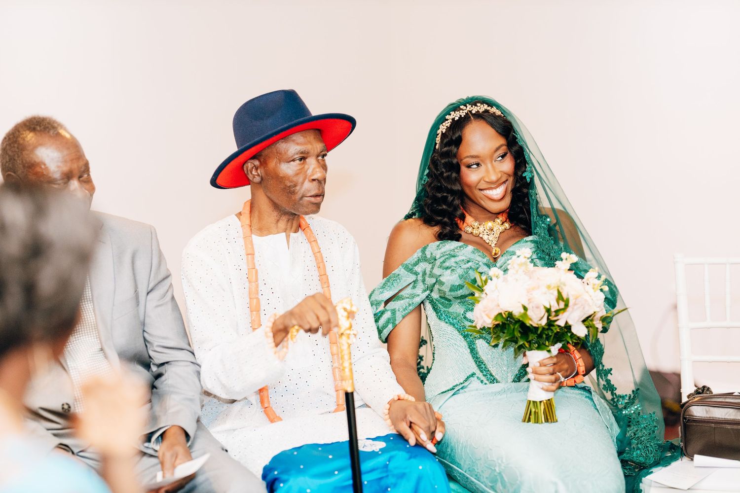 A bride in a teal traditional gown holds white flowers beside an elder in a red-brimmed hat at a cultural wedding ceremony.