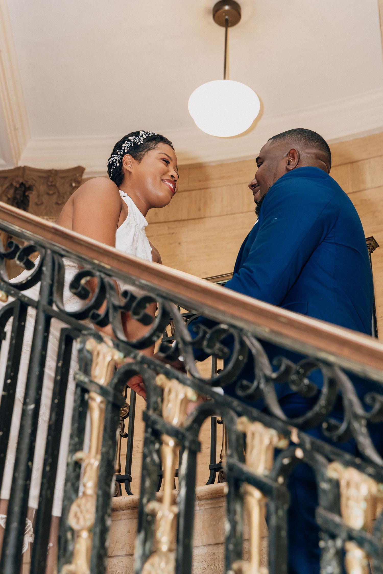 Bride in white halter dress and groom in blue suit share a joyful moment on grand staircase with iron railing.