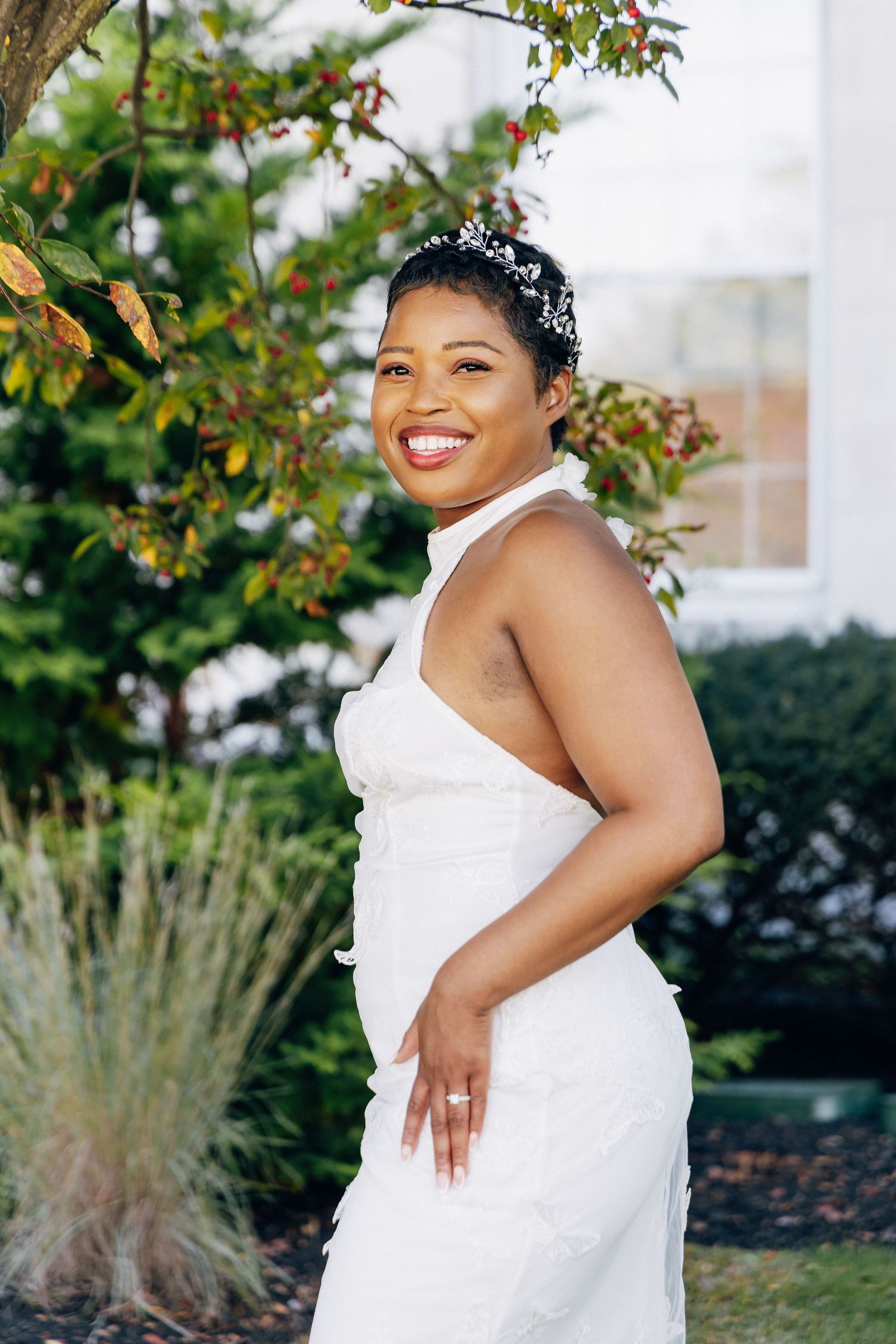 Smiling bride in white halter wedding dress with pearl headpiece posing in outdoor garden setting.