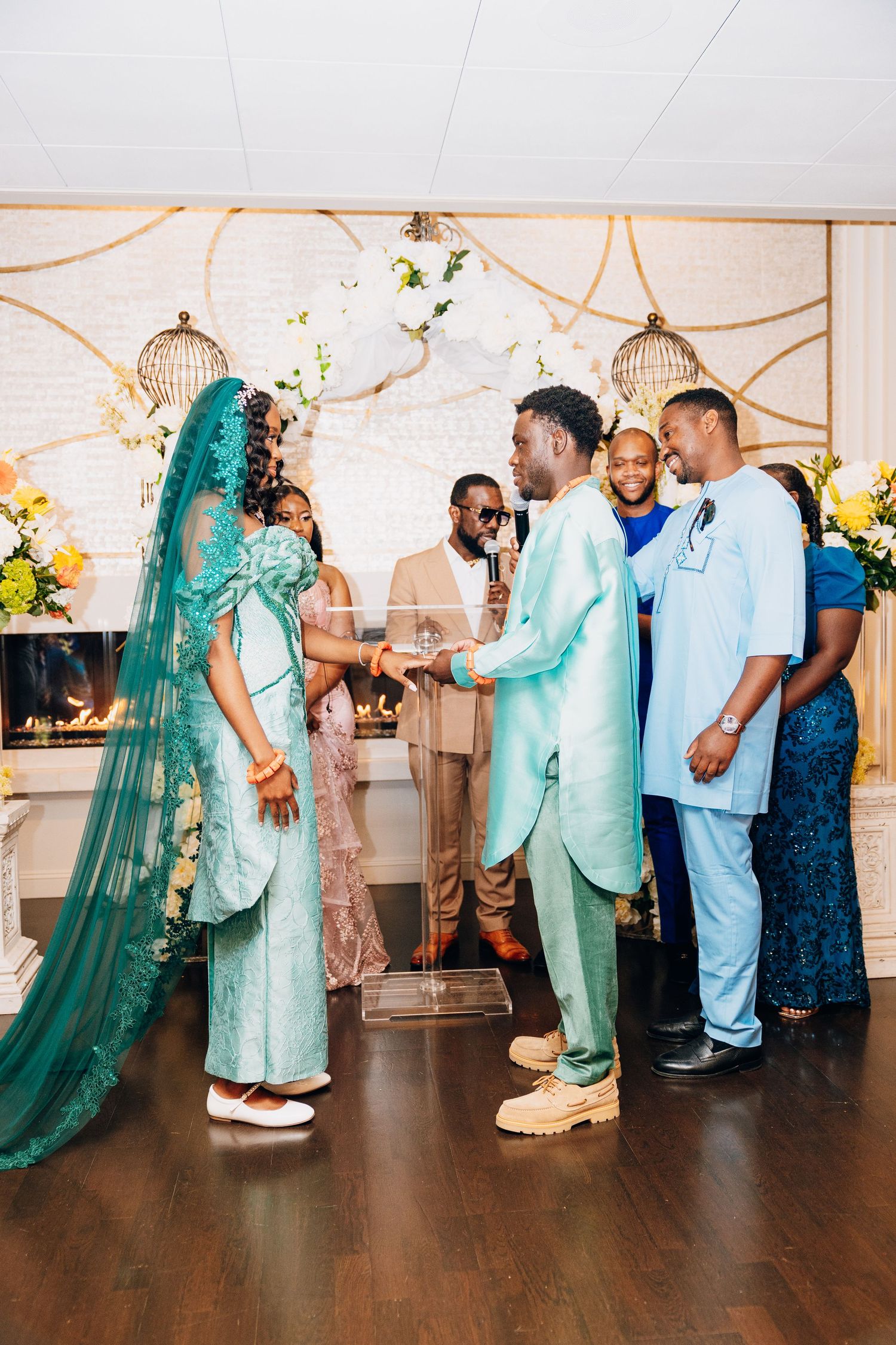 Groom in mint suit holds bride's hands during ring exchange at decorated indoor wedding ceremony.