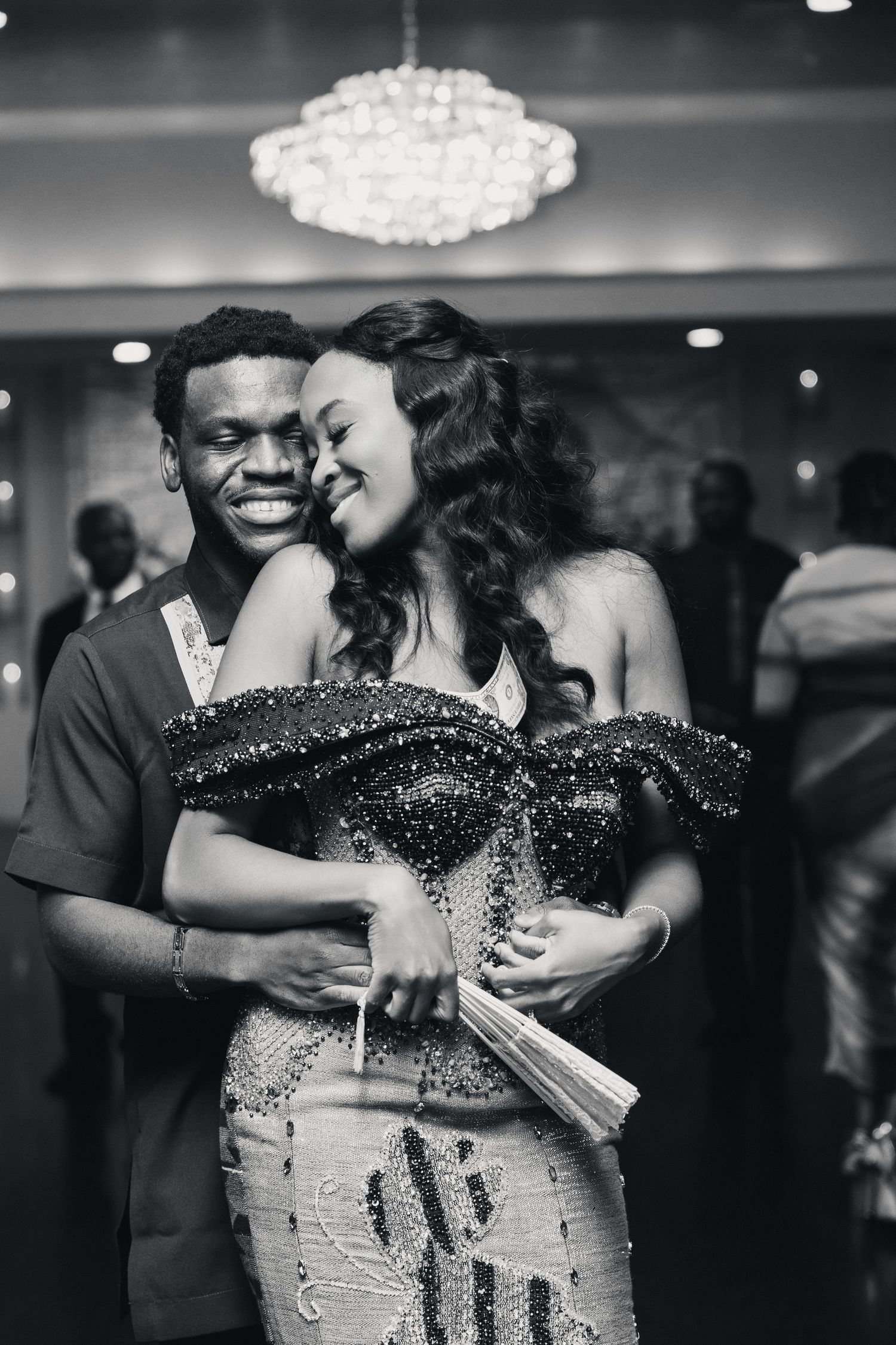 Black and white photo of a smiling couple embracing at a formal event, woman in beaded gown, chandelier backdrop.
