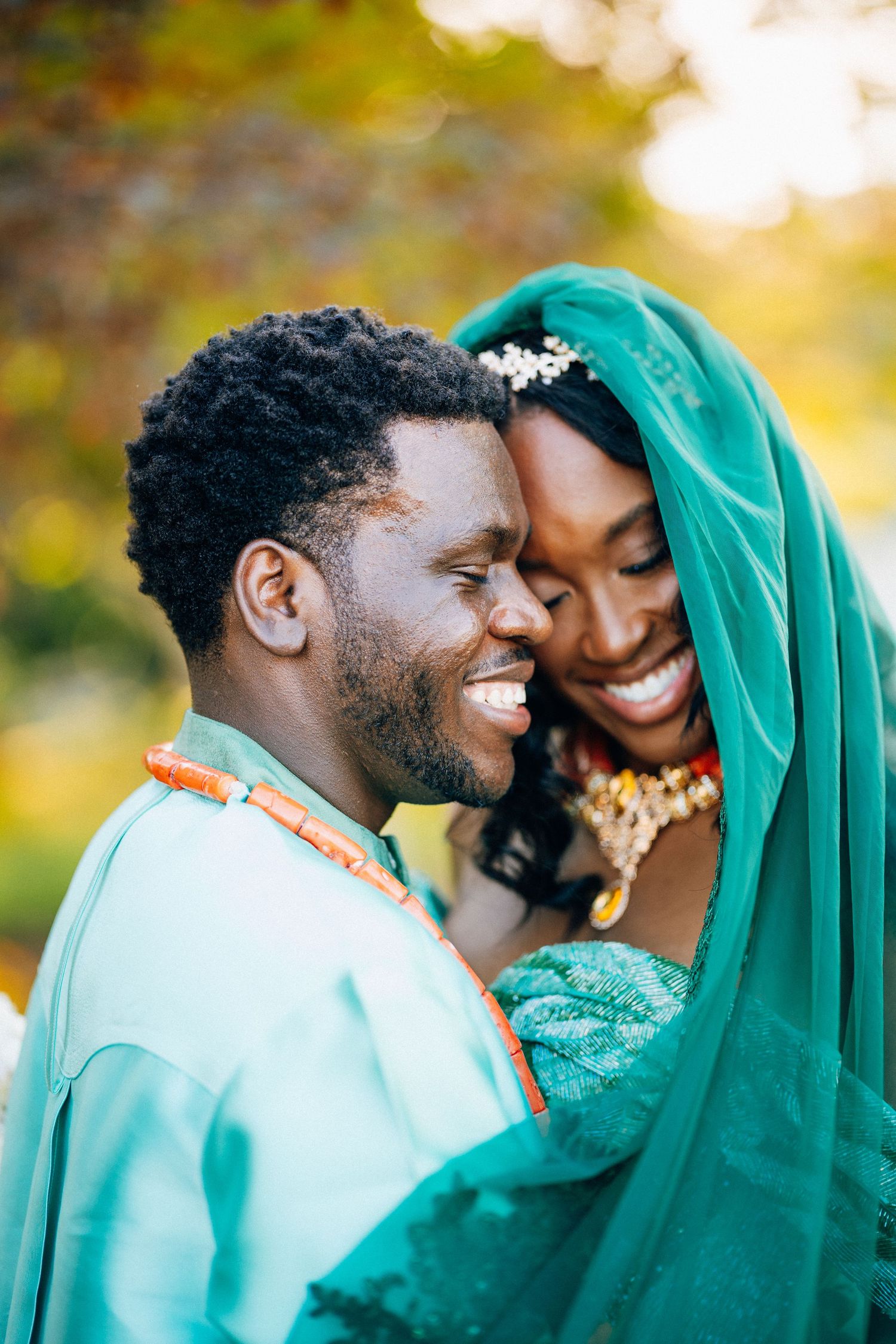 Smiling Nigerian couple in matching teal traditional wedding attire share joyful moment under veil.