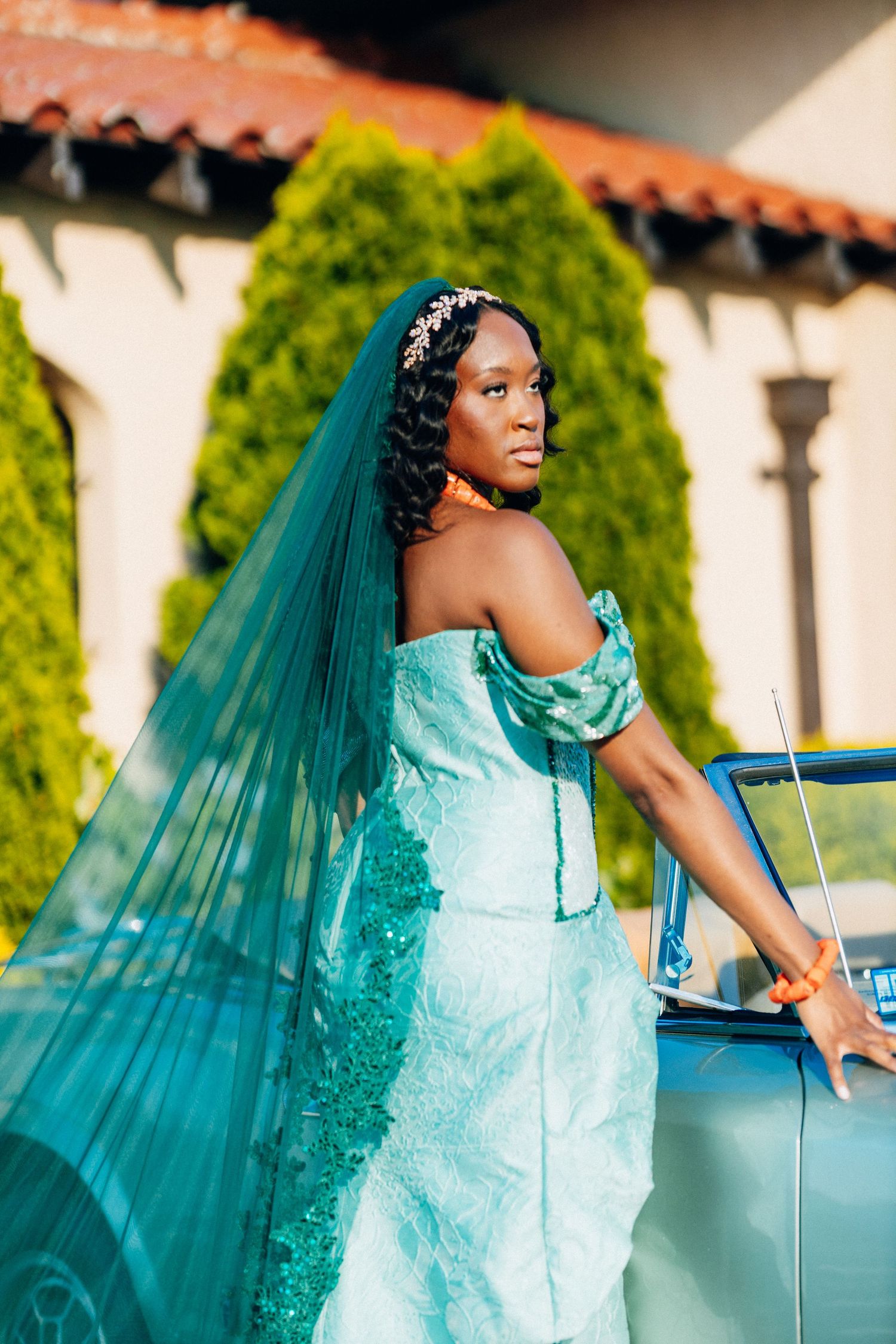 African bride in teal sequin gown and veil leans on vintage blue car.