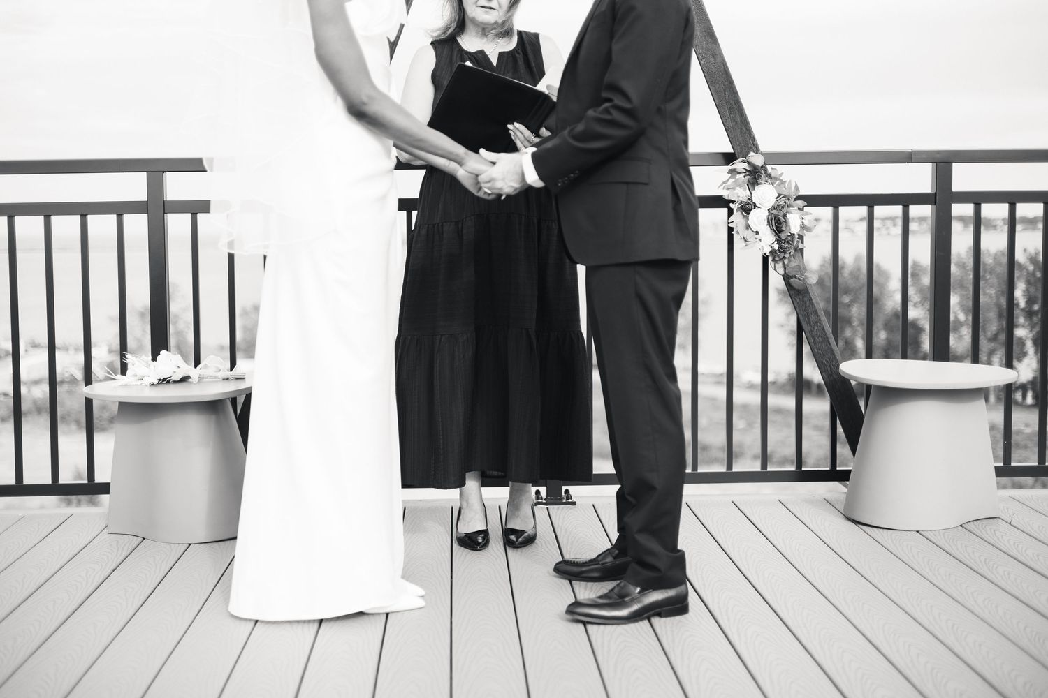Black and white photo of a couple exchanging vows with an officiant on a deck during an outdoor wedding ceremony.