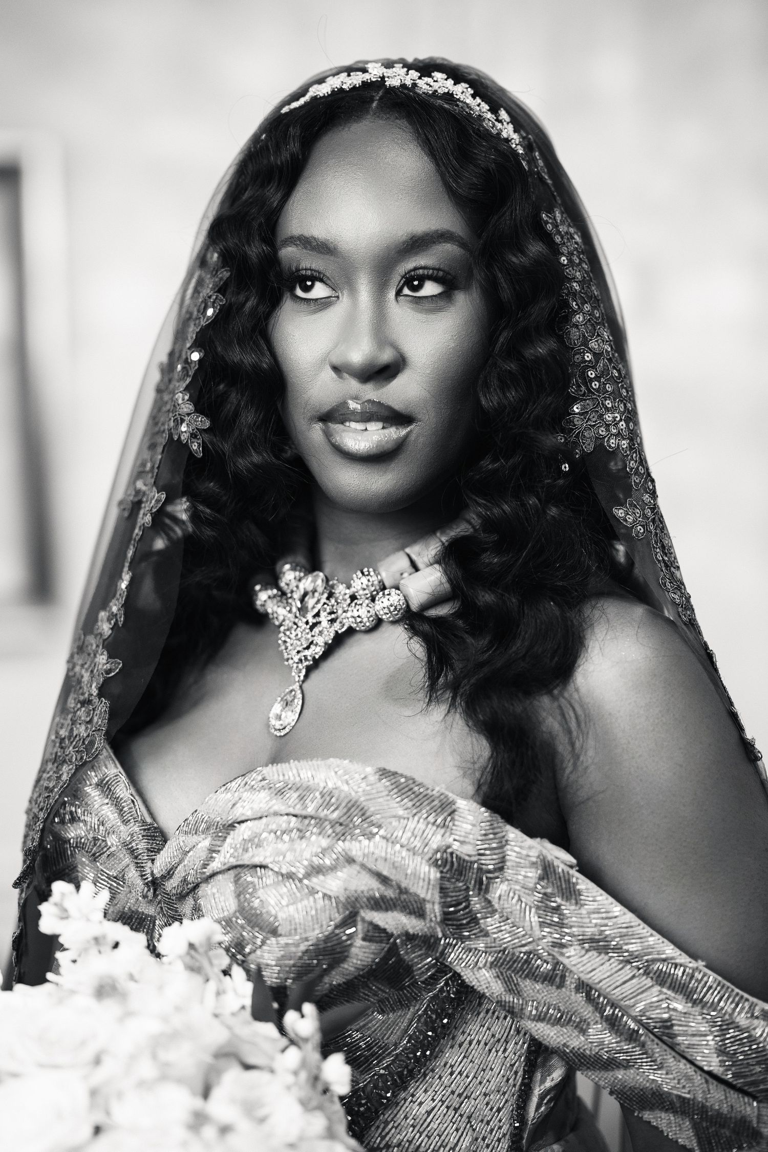 Black and white portrait of a bride in traditional attire with lace veil, floral crown, and statement necklace.