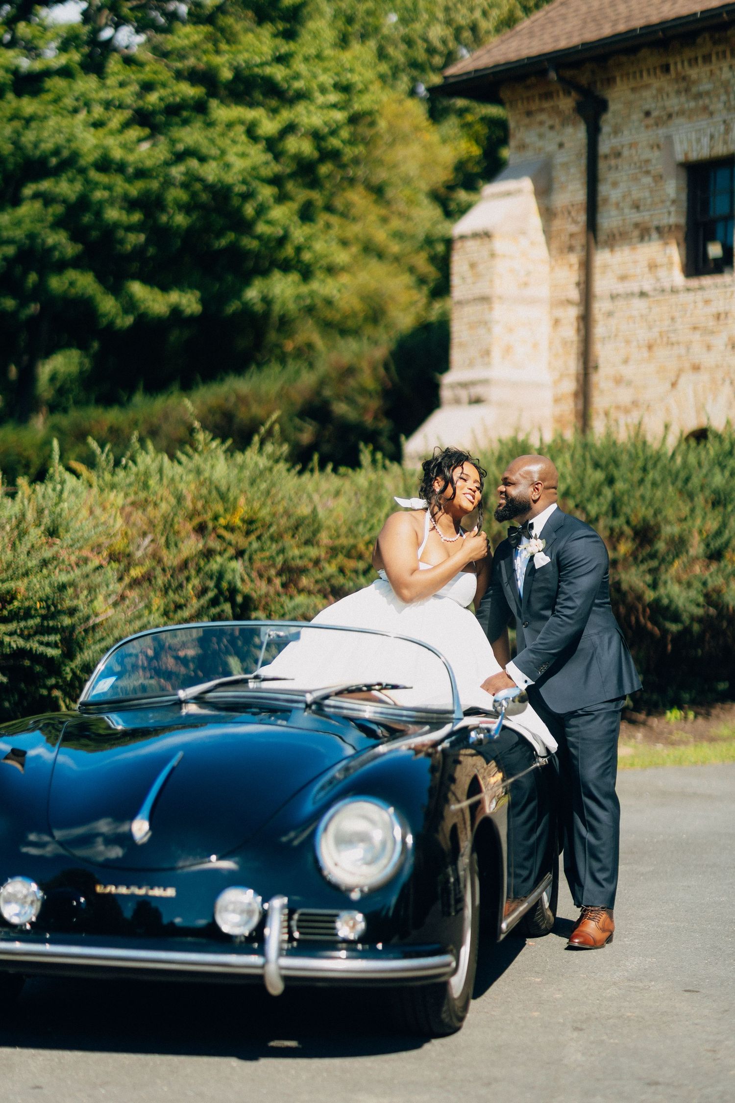 Joyful bride and groom share a laugh beside classic vintage Porsche speedster on wedding day.