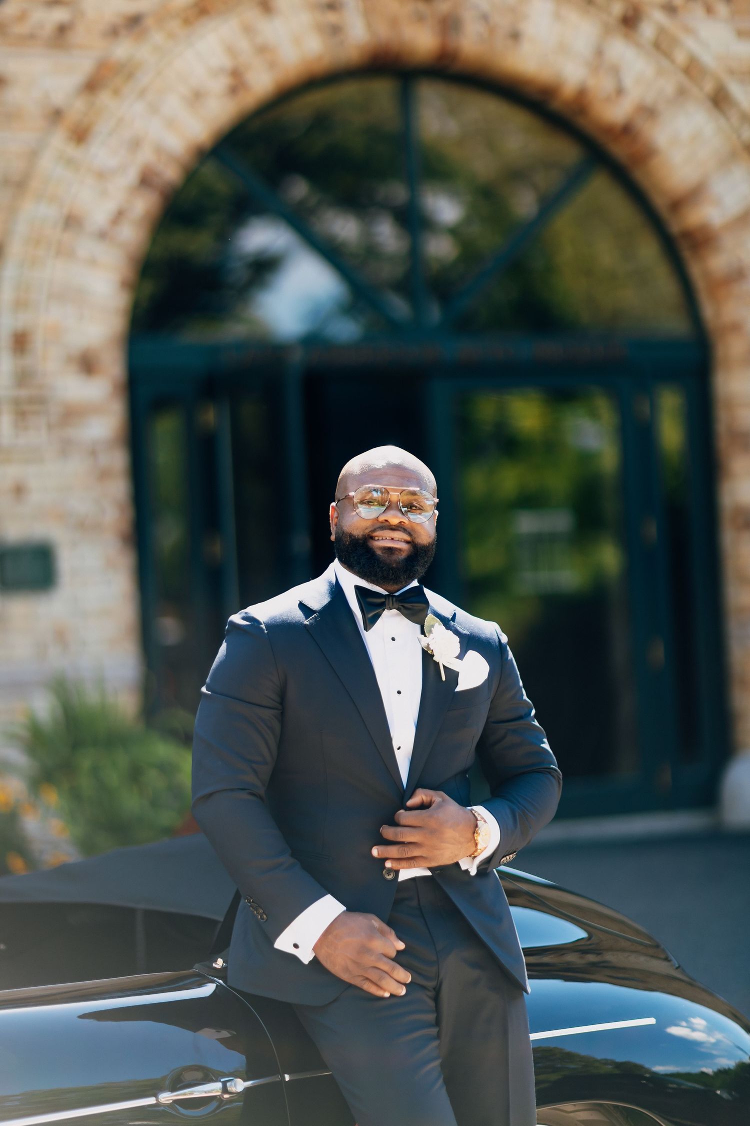 Stylish groom in navy tuxedo with boutonniere poses confidently outside elegant brick arch building.