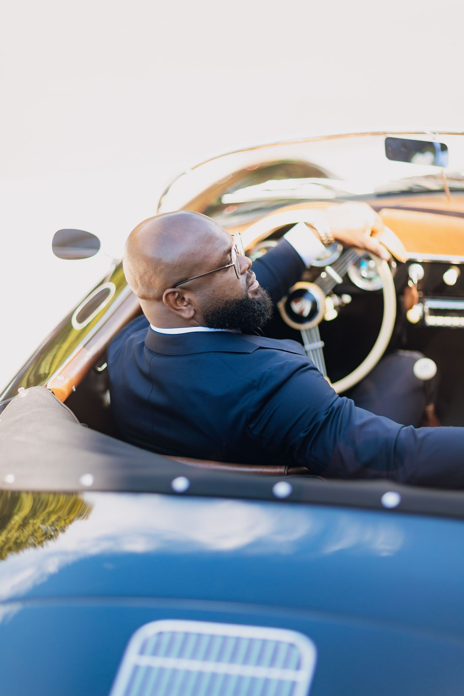 Bald groom in navy suit drives a vintage blue convertible sports car on wedding day.