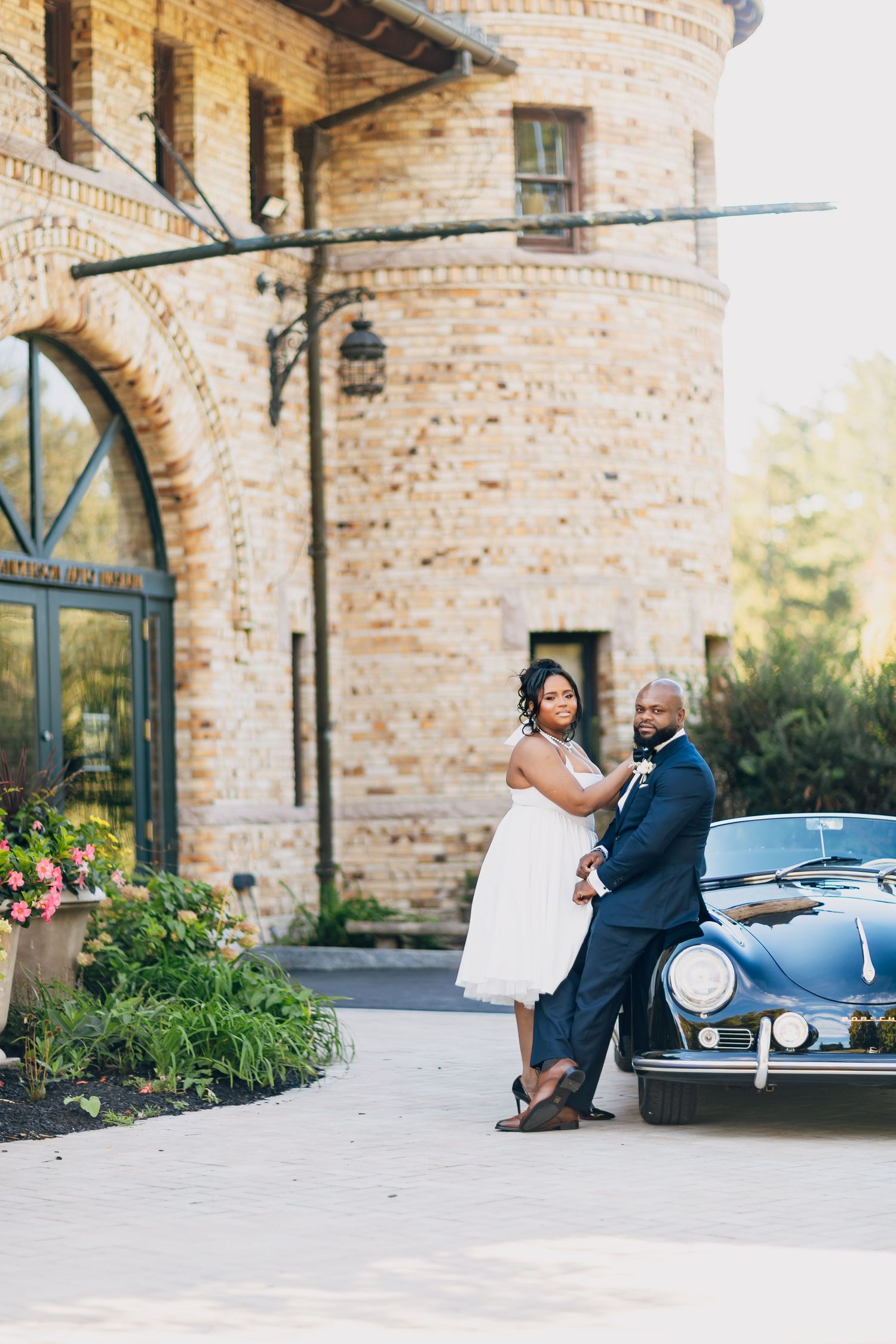 Black couple in wedding attire posing beside a vintage Porsche 356 outside a stone building.