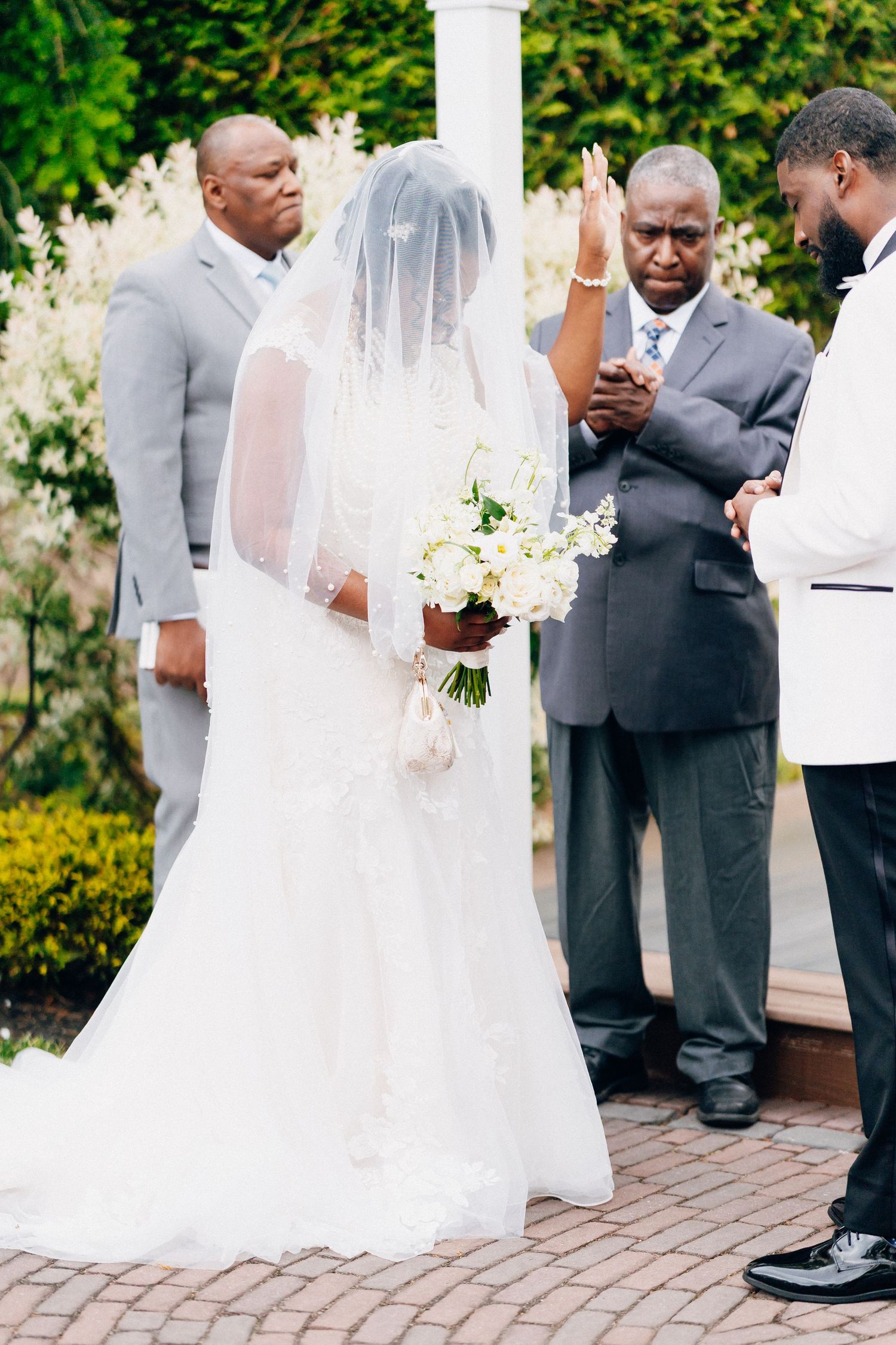 Officiant raises hand in blessing over veiled bride holding white bouquet at outdoor wedding ceremony.