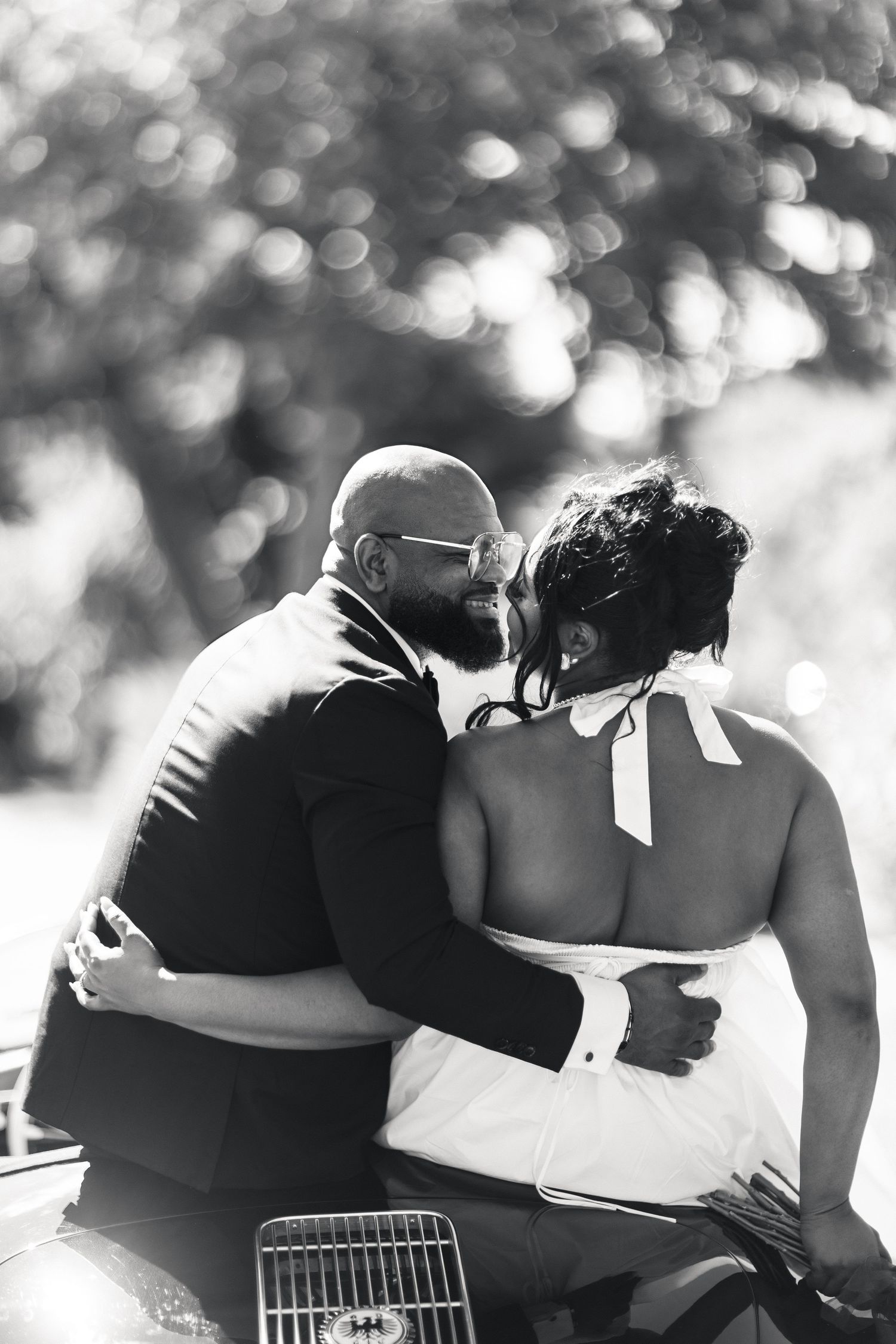 Romantic black and white photo of bride and groom embracing closely beside a vintage classic car.