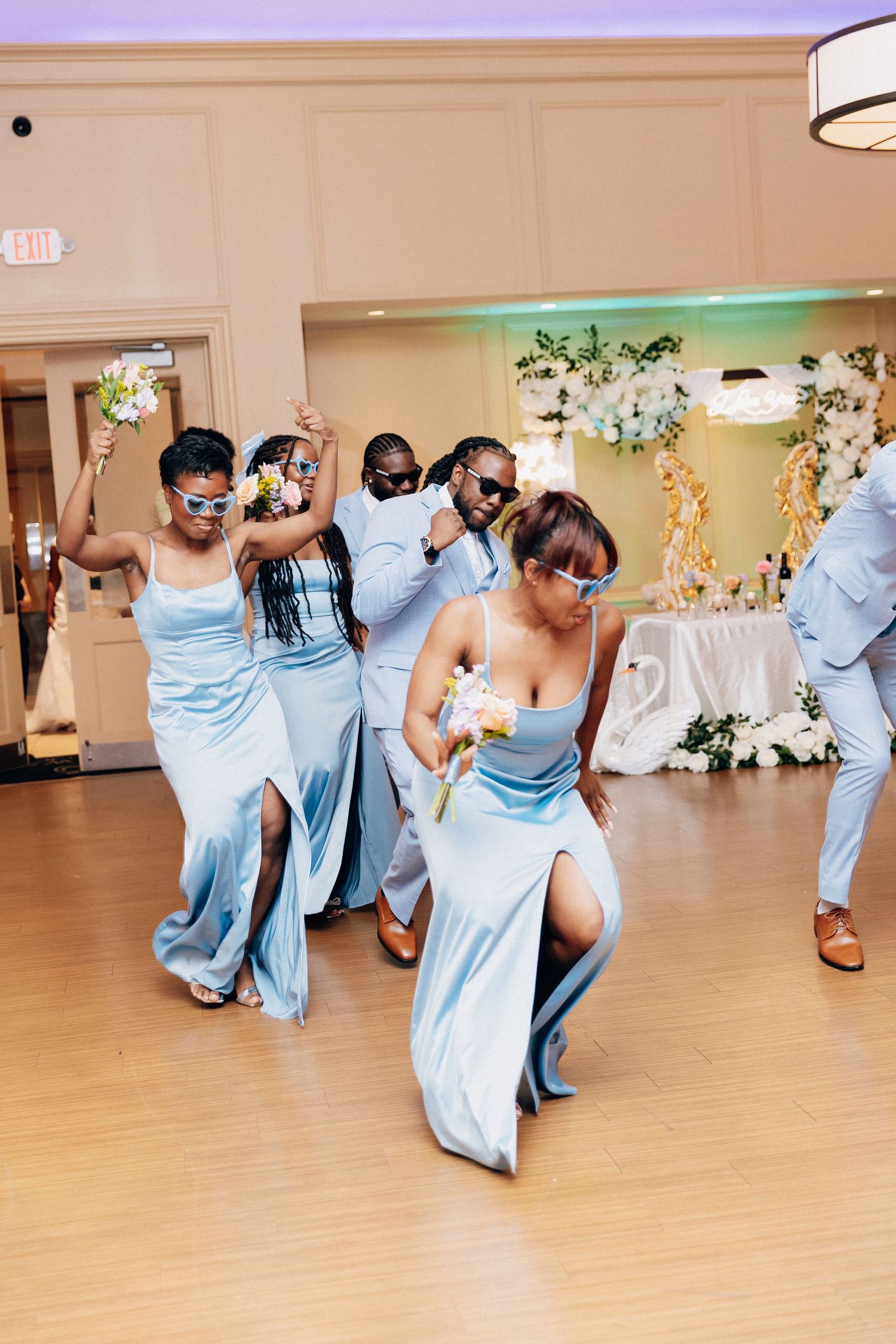 Bridesmaids in light blue satin dresses dancing energetically at a wedding reception holding white bouquets.