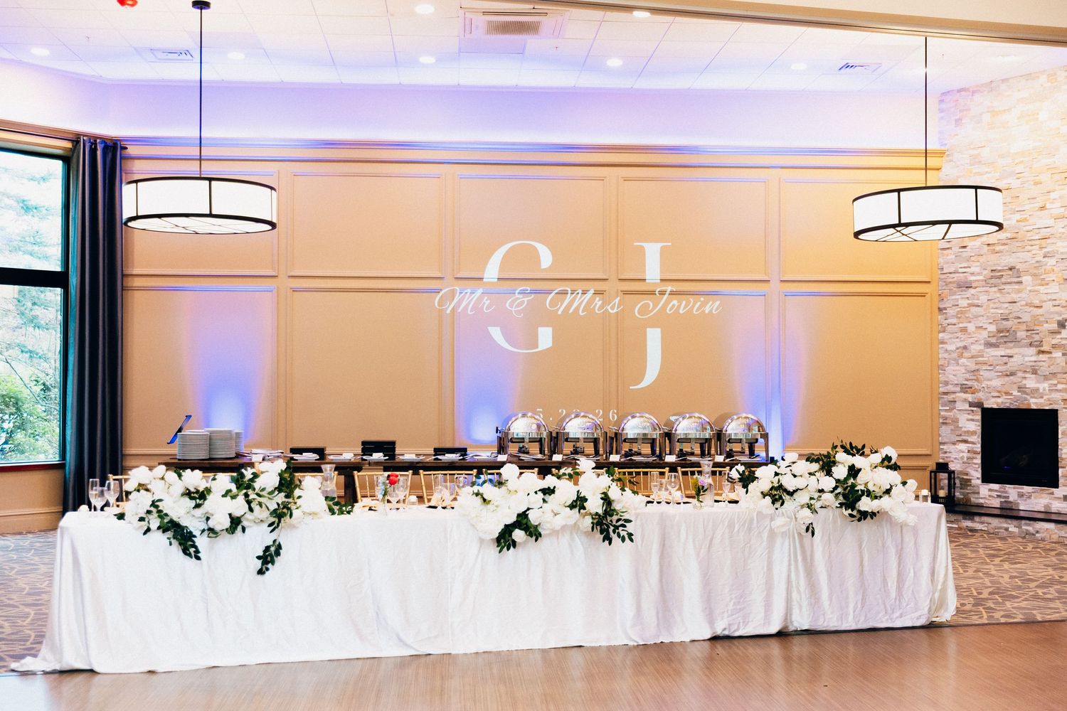 Elegant wedding reception head table with white floral arrangements and monogram backdrop lighting in a grand ballroom.