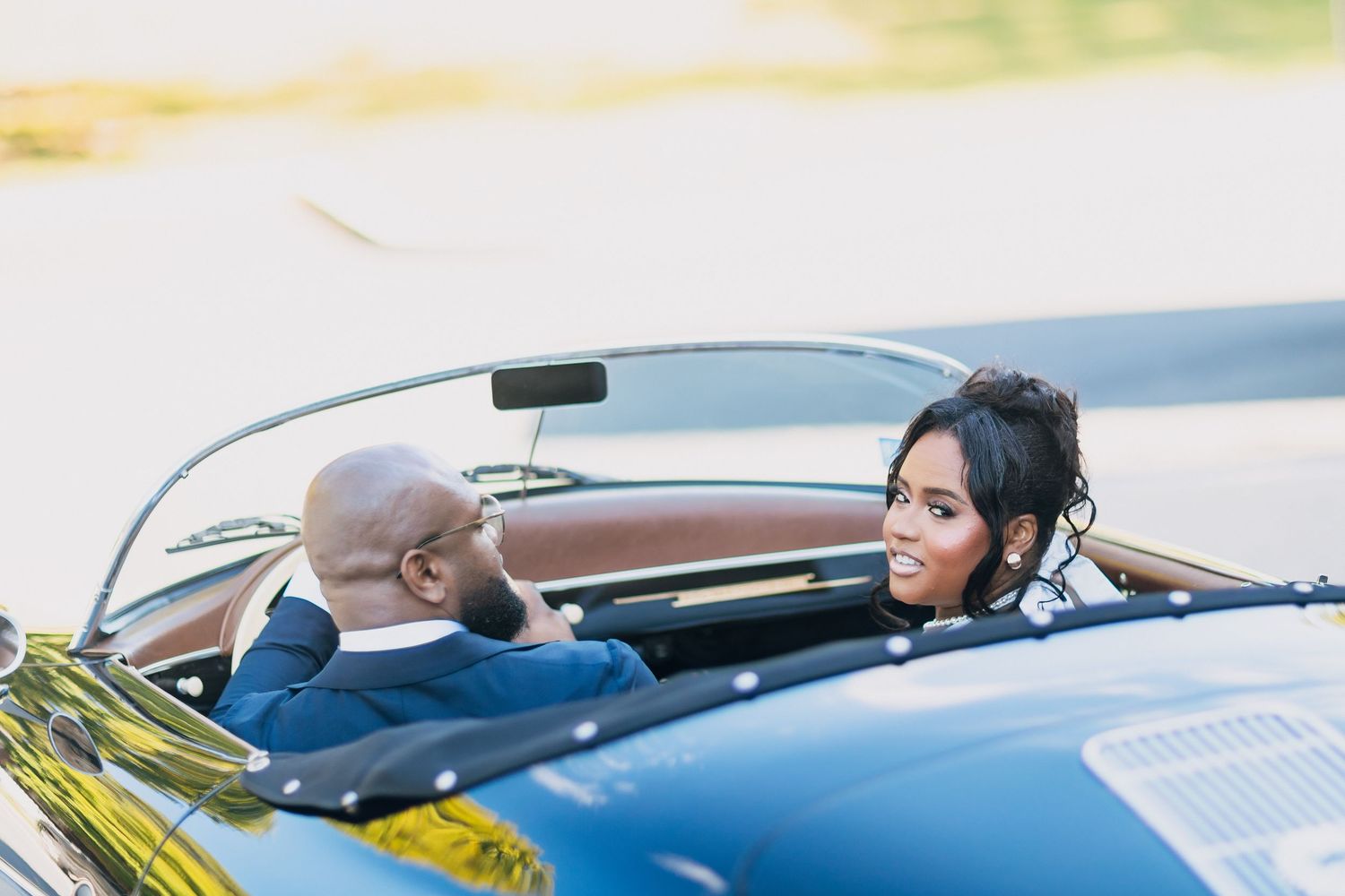 A couple sits in a vintage convertible car, dressed elegantly, sharing a romantic moment on a sunny day.