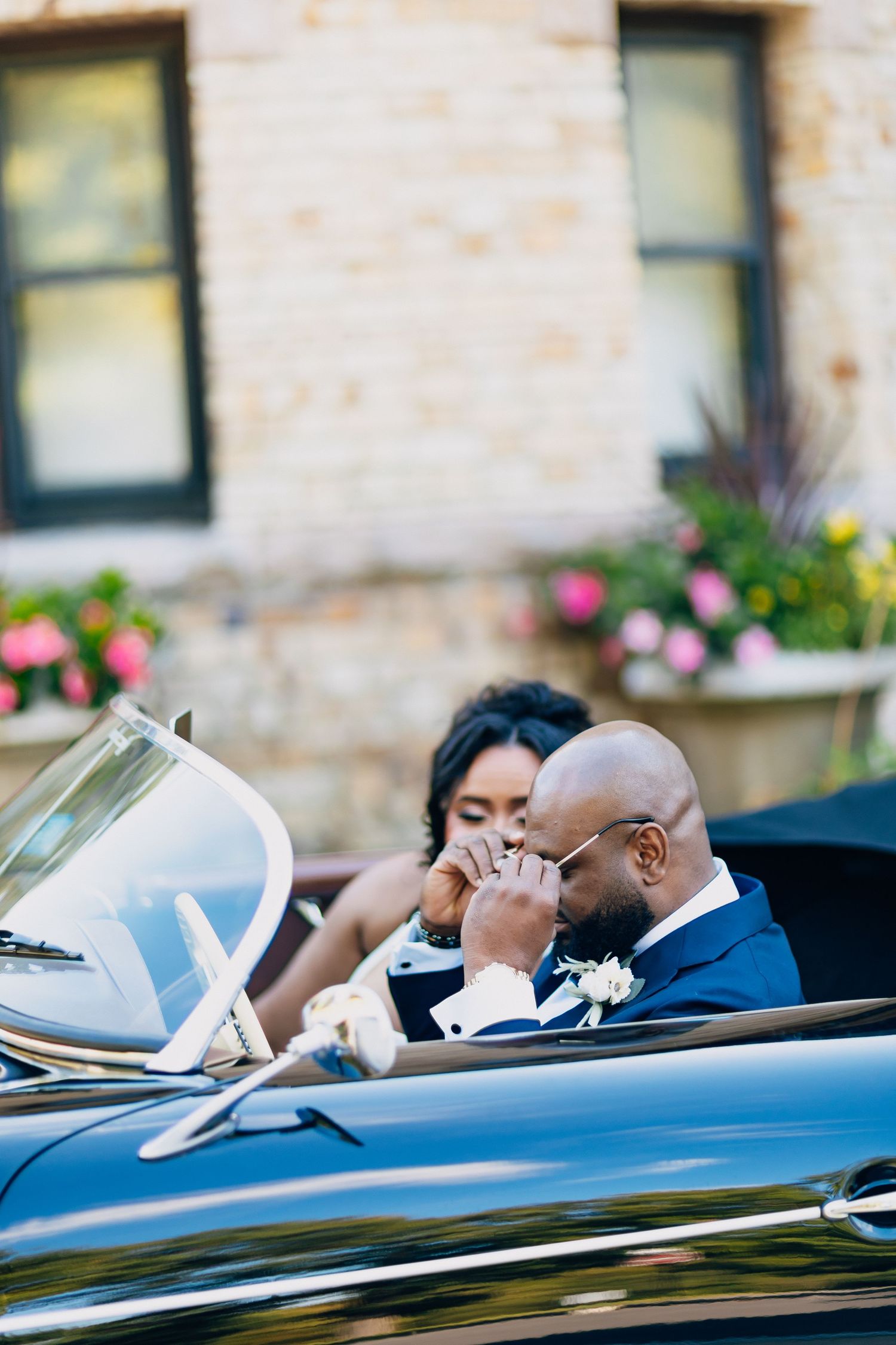 Groom in navy suit kisses bride's hand while seated together in a vintage black convertible.