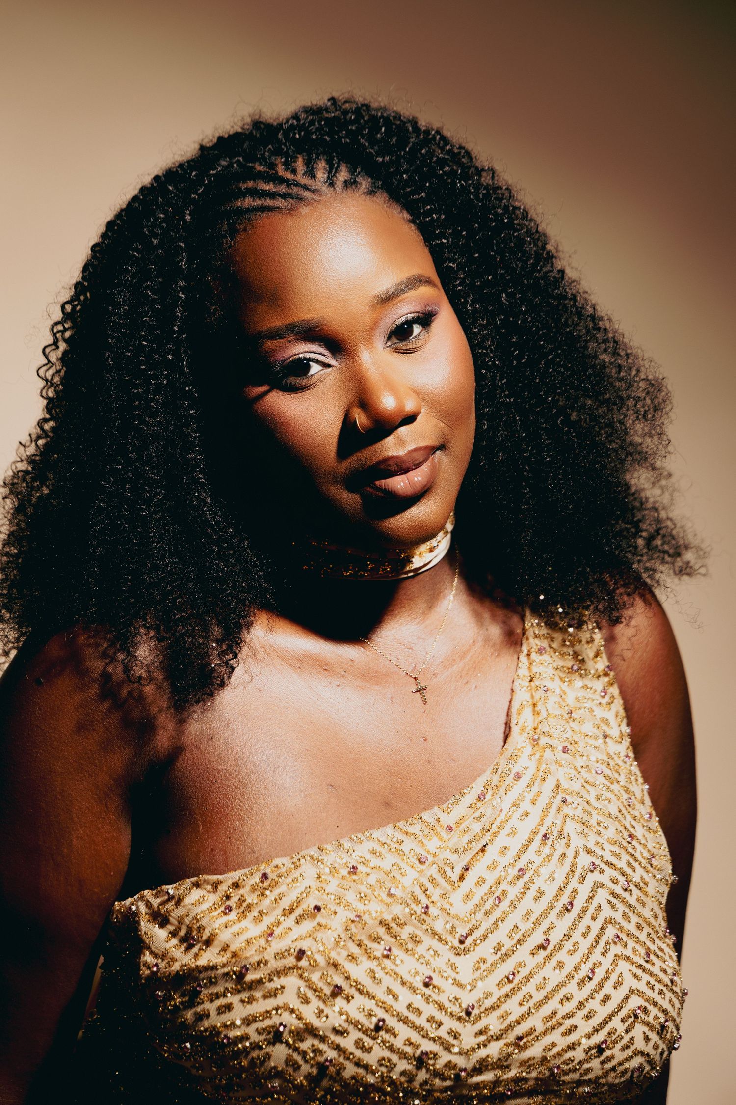 A young Black woman with voluminous curly braided hair wearing a gold sequined one-shoulder dress poses confidently.