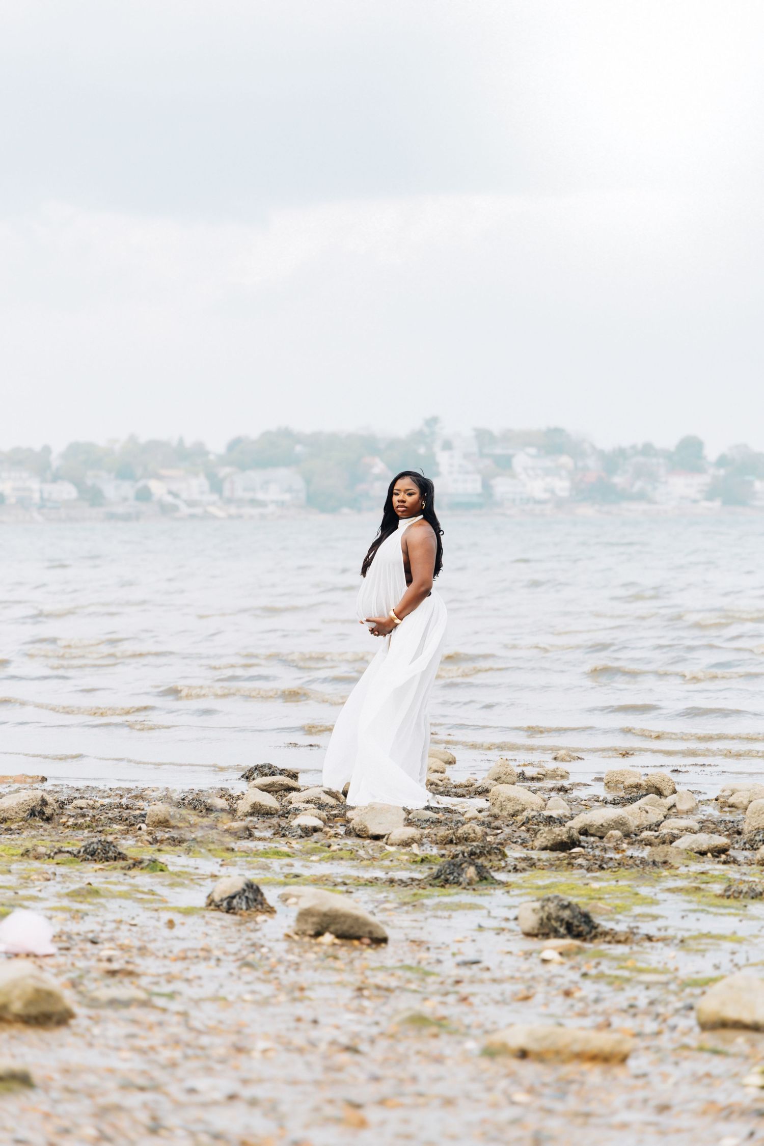 Pregnant woman in white dress standing on rocky shoreline with ocean and houses in background.