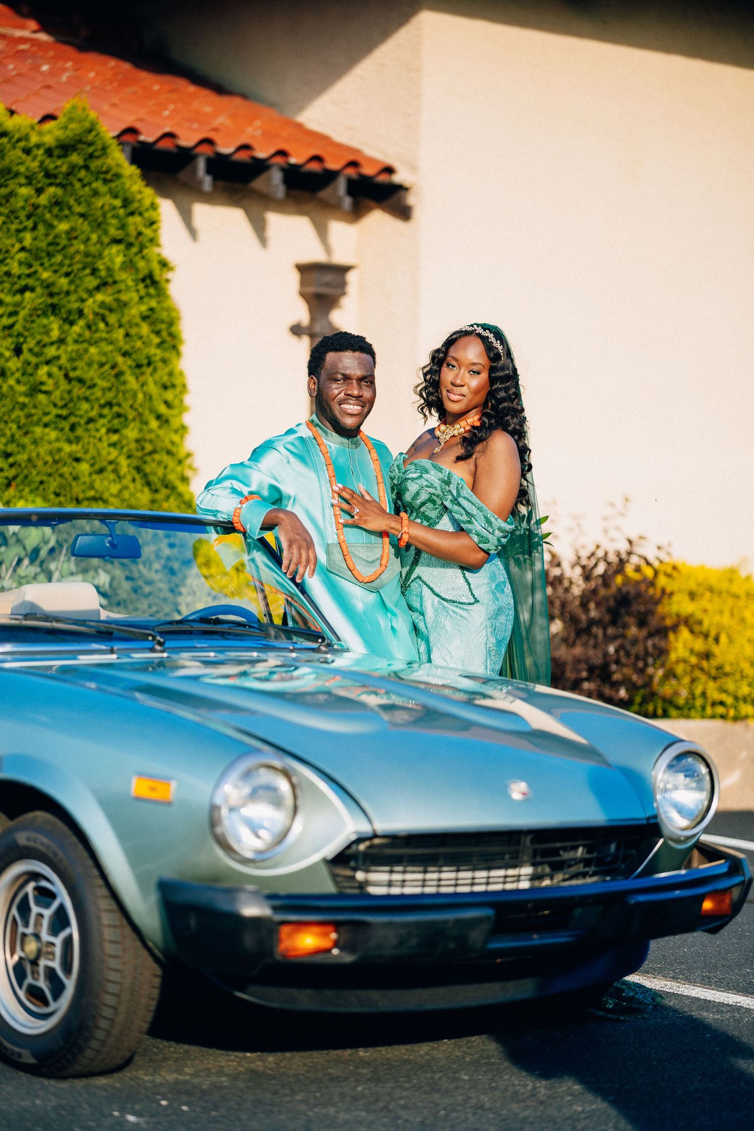 Couple in matching teal African attire posing by a classic blue convertible sports car outside a building.