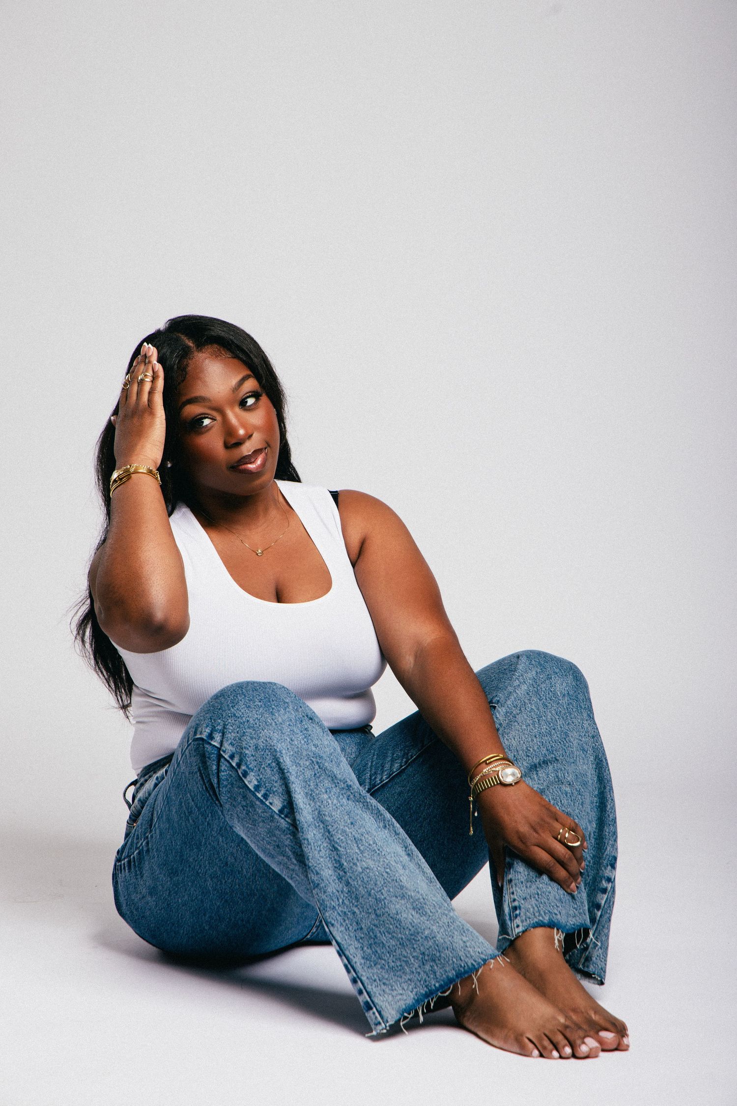 A young Black woman sits on a white floor in blue jeans and white tank top, hand raised to her head.