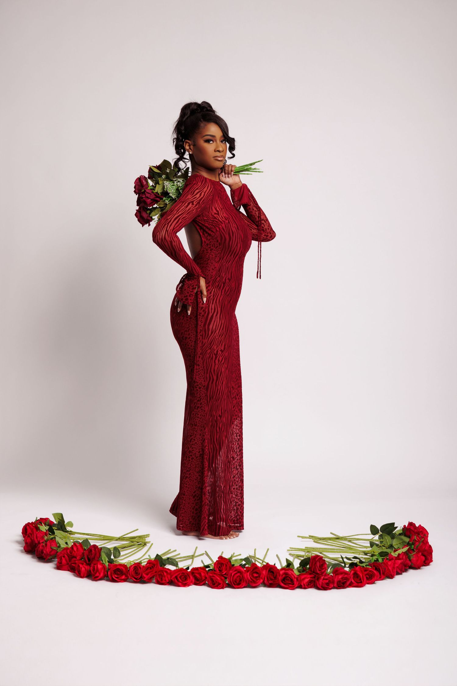 Woman in red sequin gown holding roses, surrounded by red roses arranged in arc on white background.