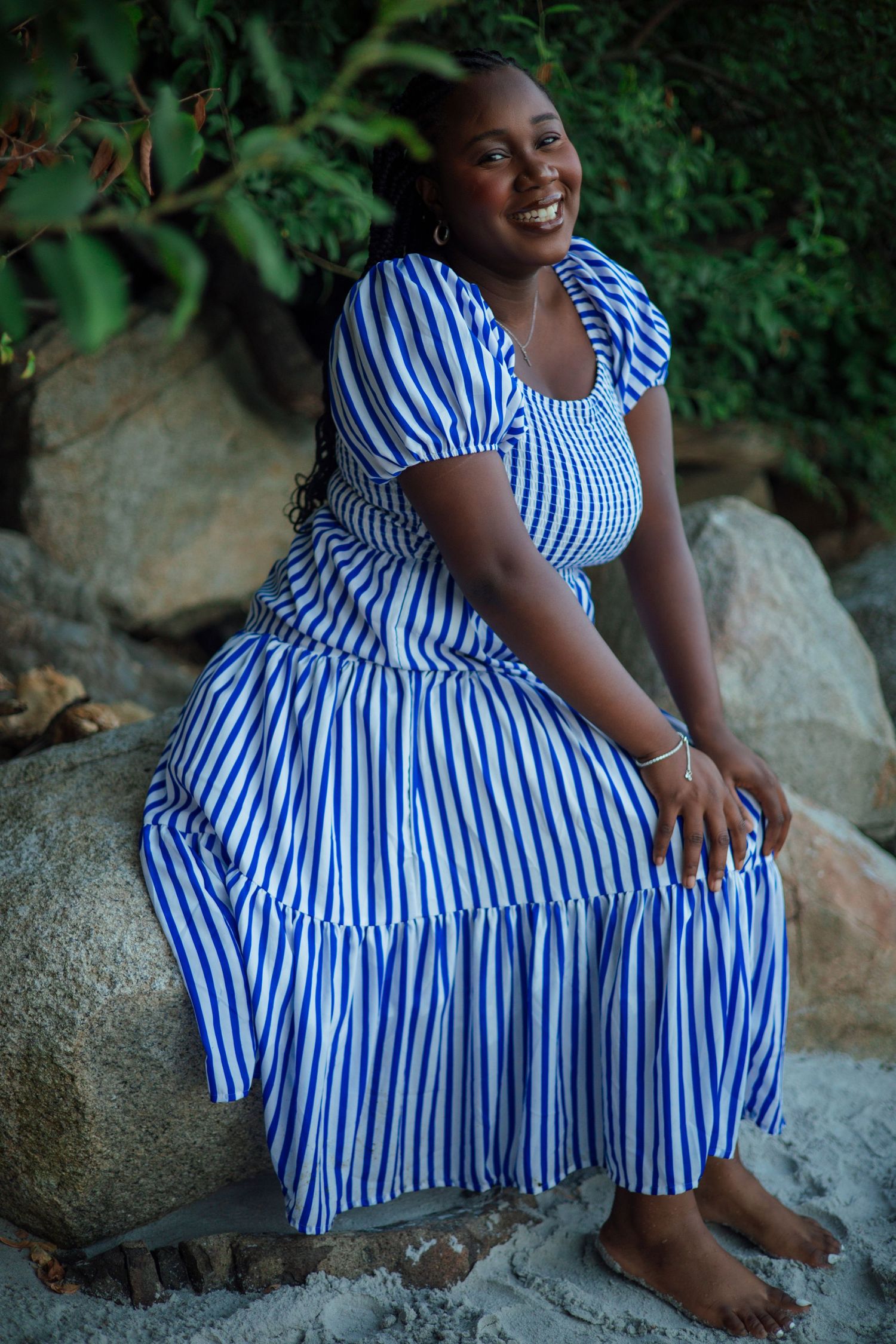 Smiling woman in blue and white striped maxi dress sitting on rocks outdoors with green foliage background.