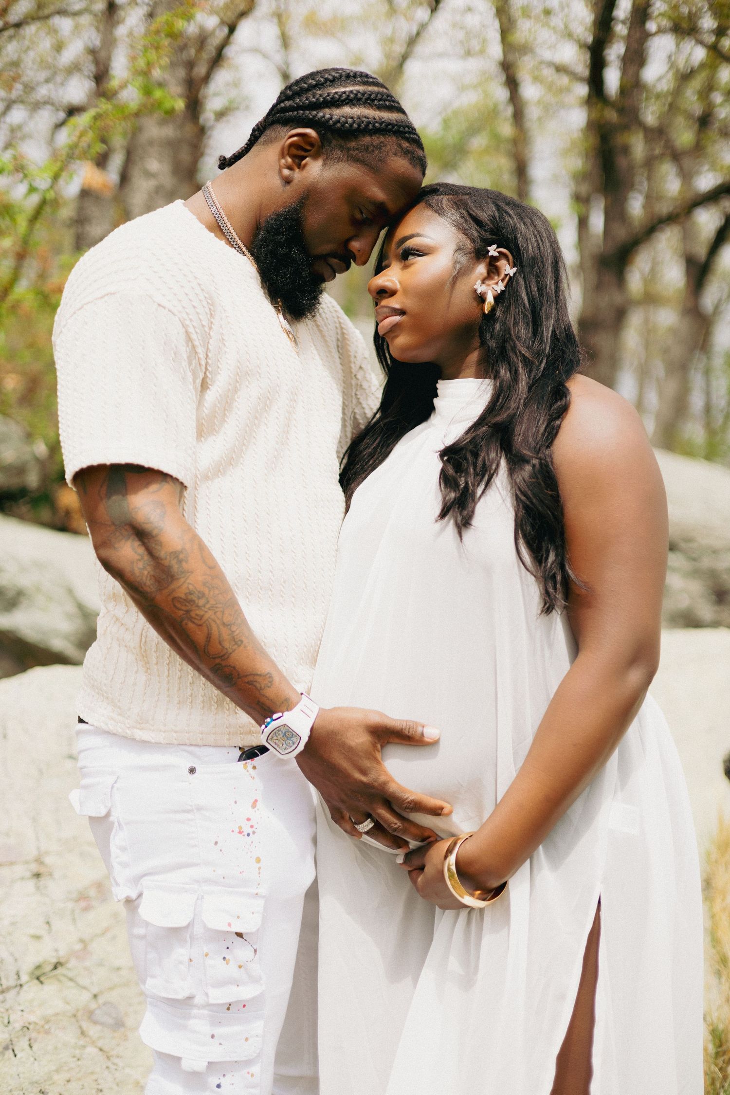 A couple in matching white outfits share a tender moment outdoors, with the woman visibly pregnant.