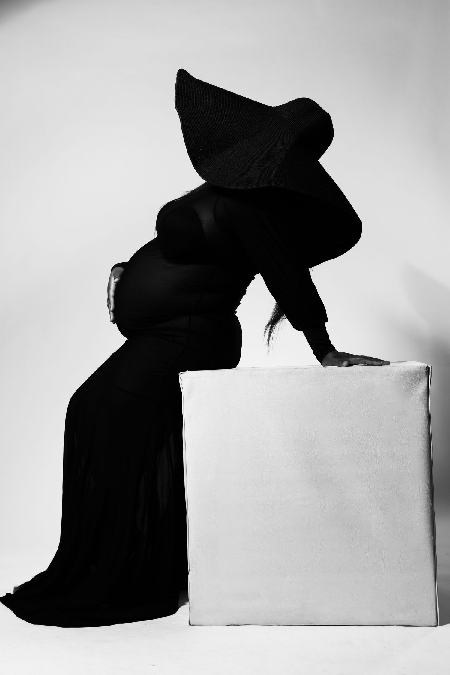 Black and white photo of a pregnant woman in a dramatic black gown and hood, leaning over a white cube.