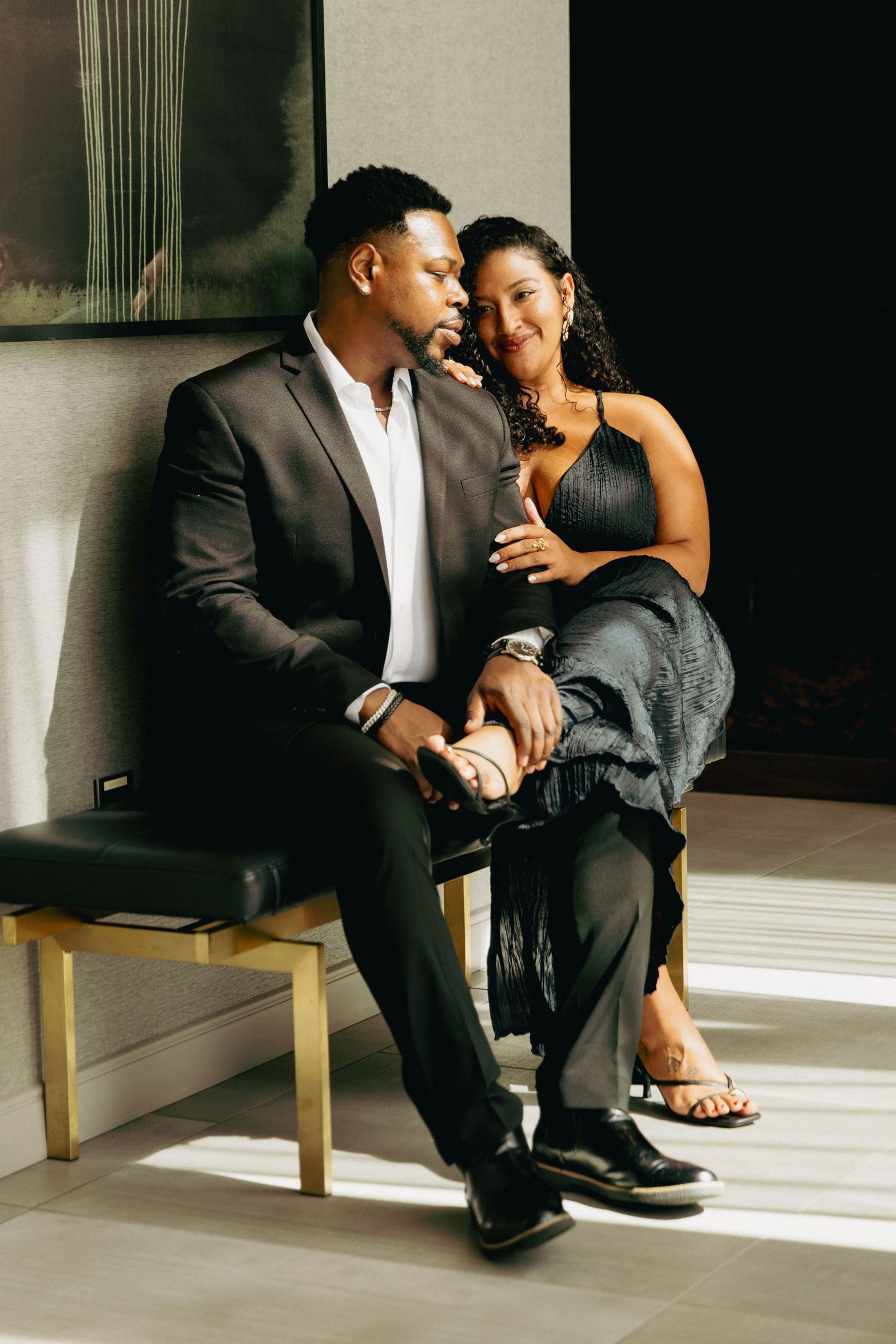 Elegant couple in formal attire sitting together on a bench, sharing an intimate moment in a sunlit room.