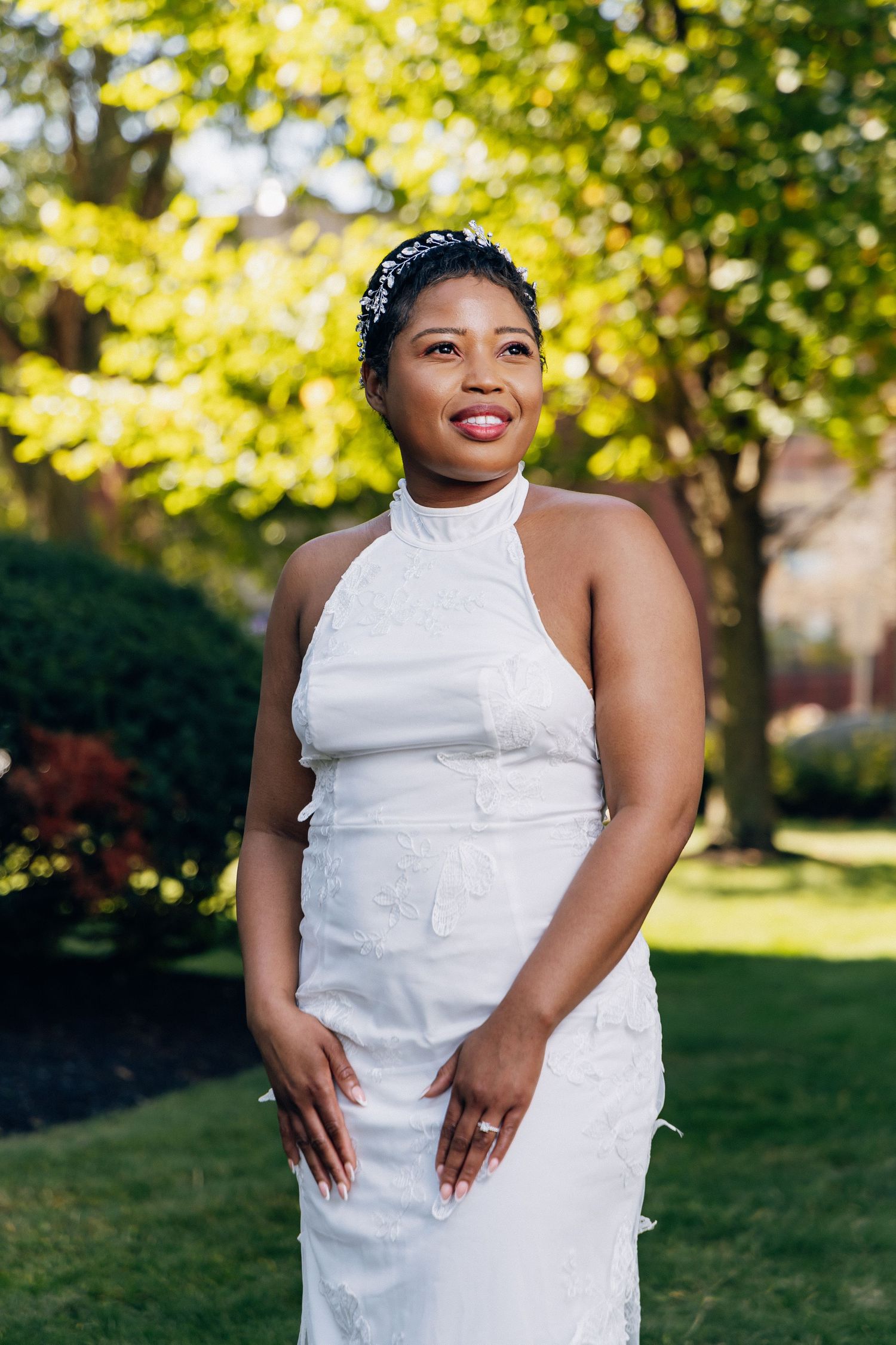 Smiling bride in white halter wedding gown with floral details poses outdoors in lush green garden.