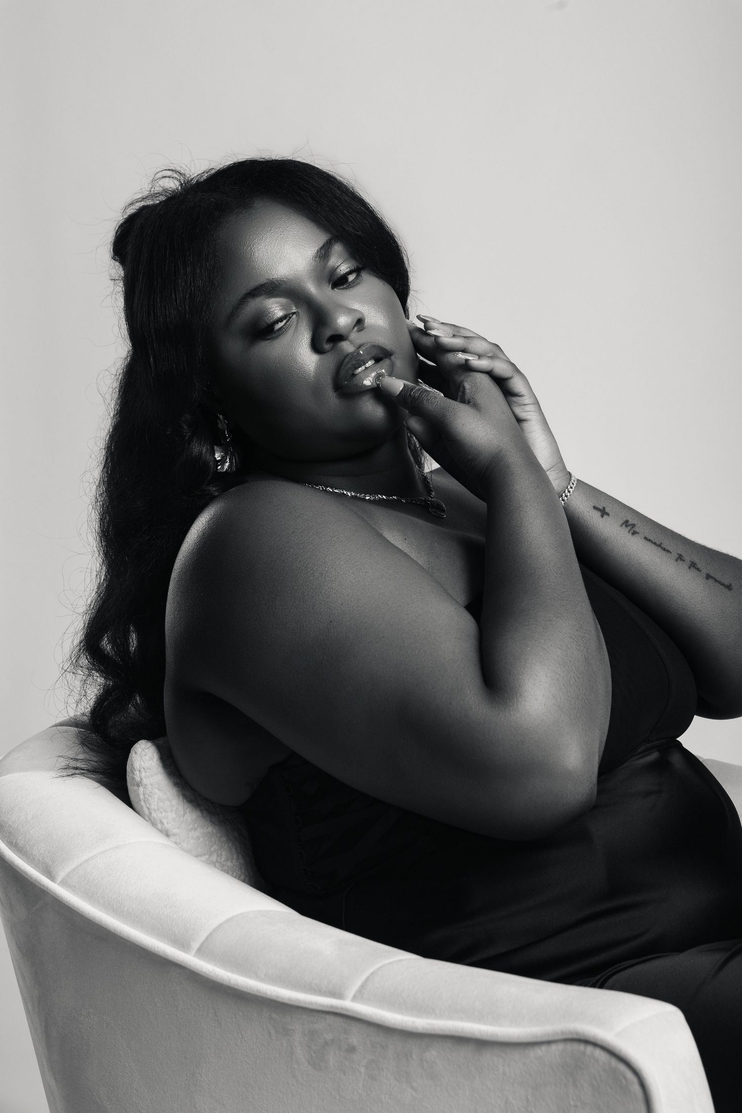 Black and white portrait of a woman sitting in a white chair, posing thoughtfully with hand near her face.