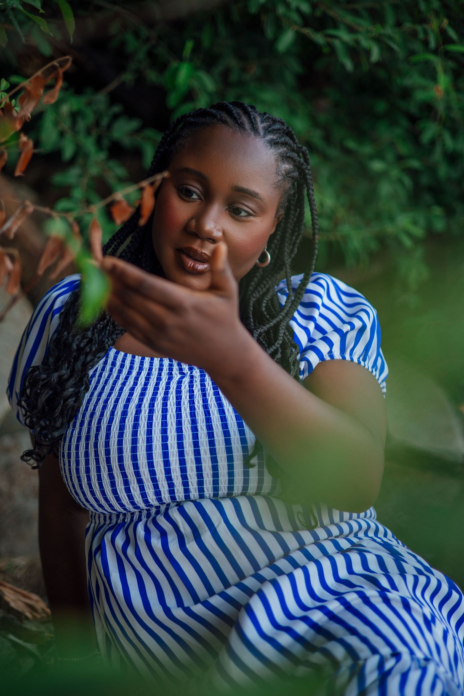 A young Black woman in a blue striped dress sits outdoors, reaching toward a green leaf among lush garden foliage.