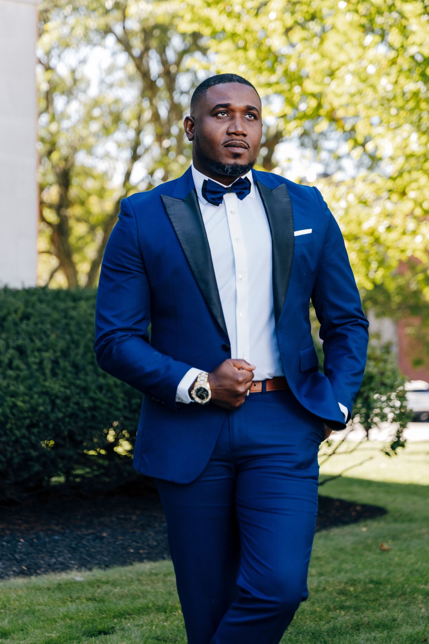 A groom in a navy blue tuxedo gazes thoughtfully while standing in a lush outdoor garden setting.