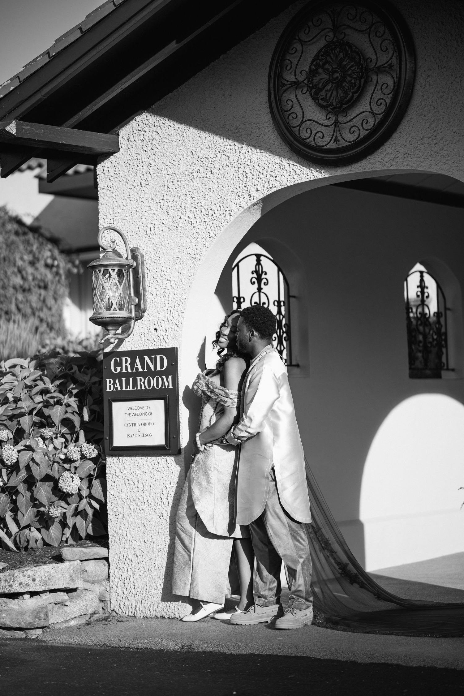 Black and white photo of newlywed couple embracing outside Grand Ballroom venue in traditional Nigerian wedding attire.