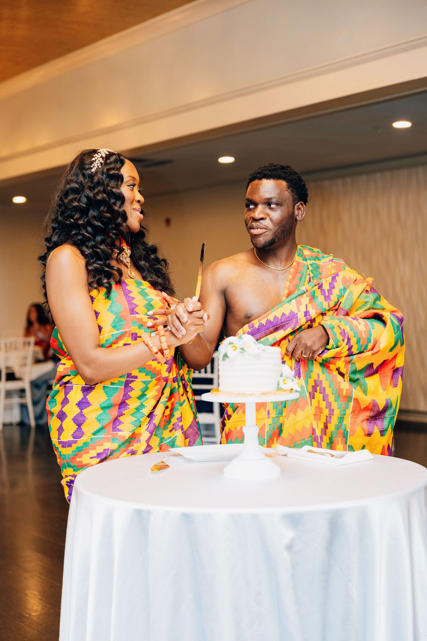 Couple in vibrant Kente cloth attire cutting their wedding cake at a traditional Ghanaian wedding reception.