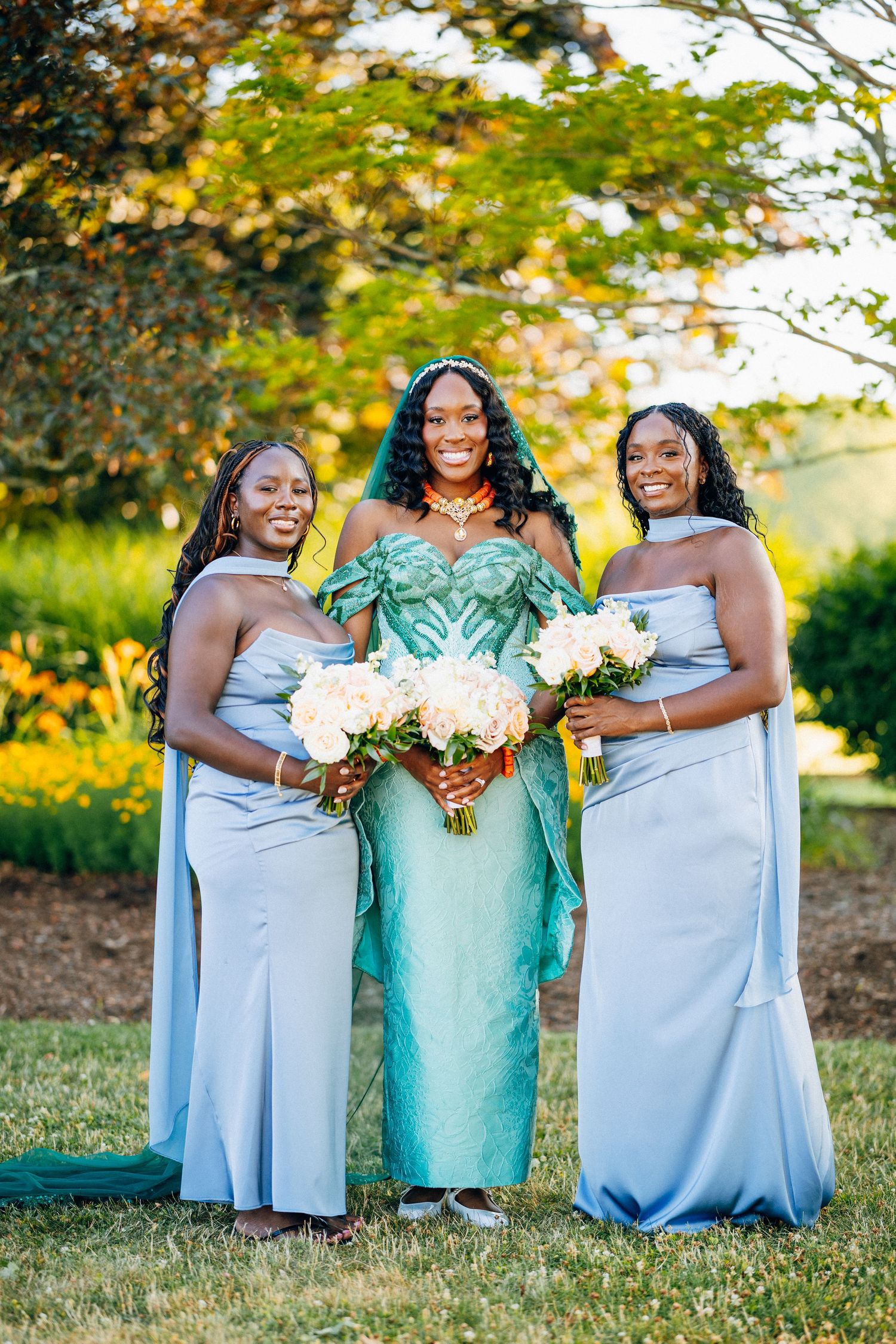 Three women in elegant gowns pose outdoors, two in light blue bridesmaid dresses and one in a green beaded bridal gown.