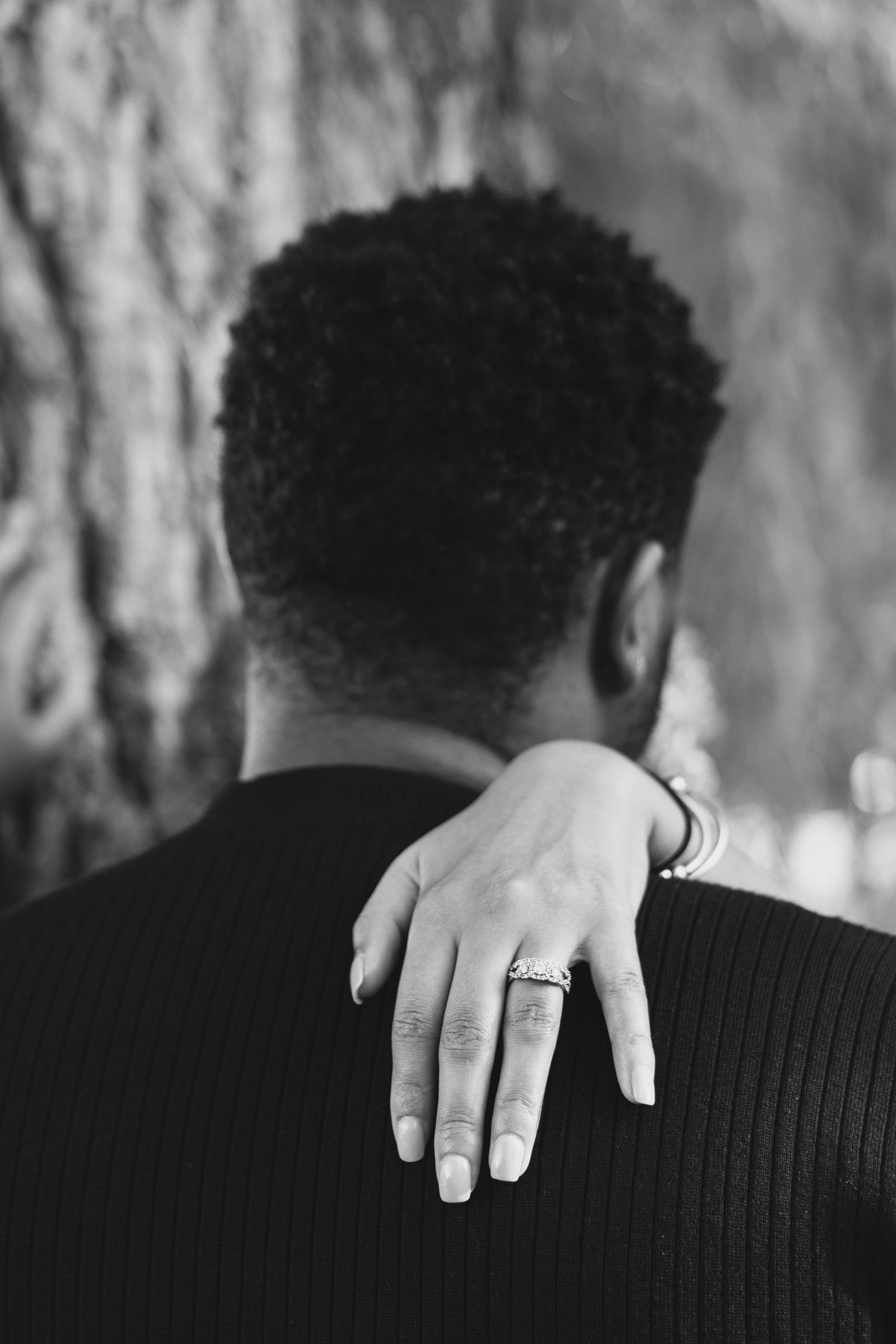 Close-up black and white photo of a woman's hand with engagement ring on man's shoulder.